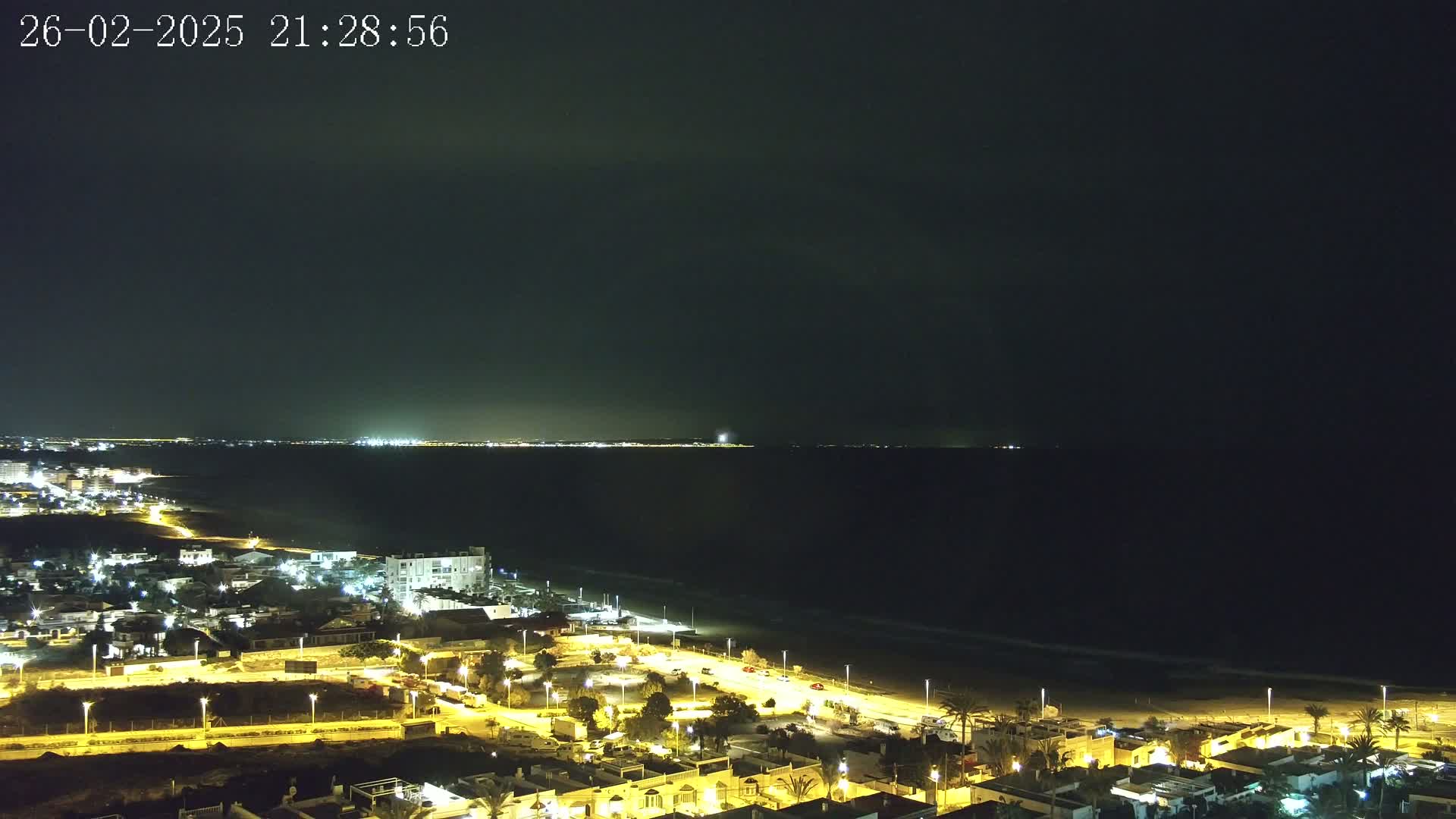 A nighttime coastal scene shows a dark sea under a mostly clear sky, with a brightly lit town along the shore and distant lights on the opposite coast.