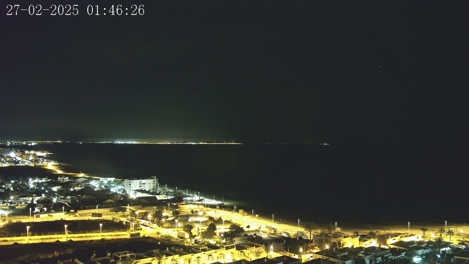 A nighttime, aerial view shows a coastal town illuminated by streetlights, next to a dark ocean with a faintly lit distant shoreline under a clear, dark sky.