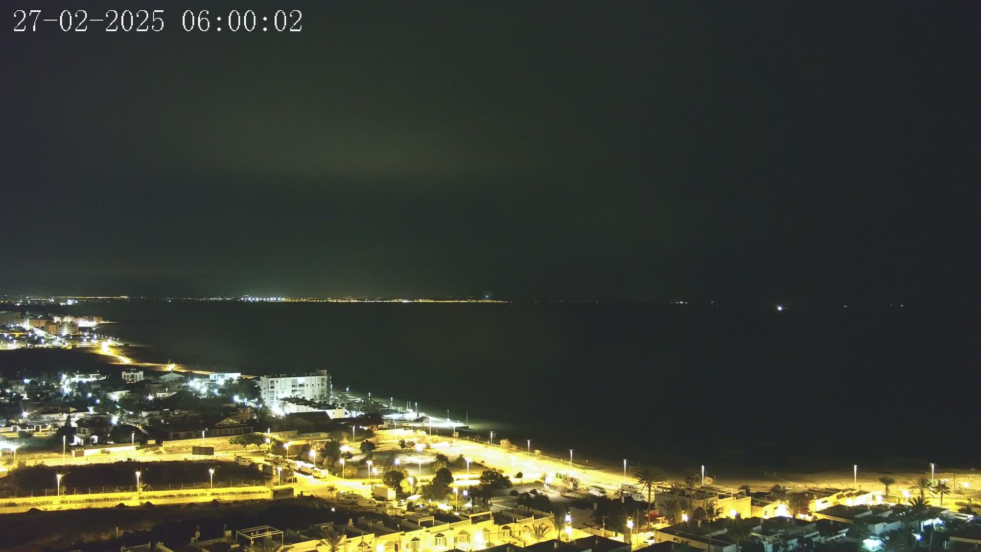 A nighttime, high-angle view shows a dark sea with a brightly lit coastline featuring residential buildings and streets.