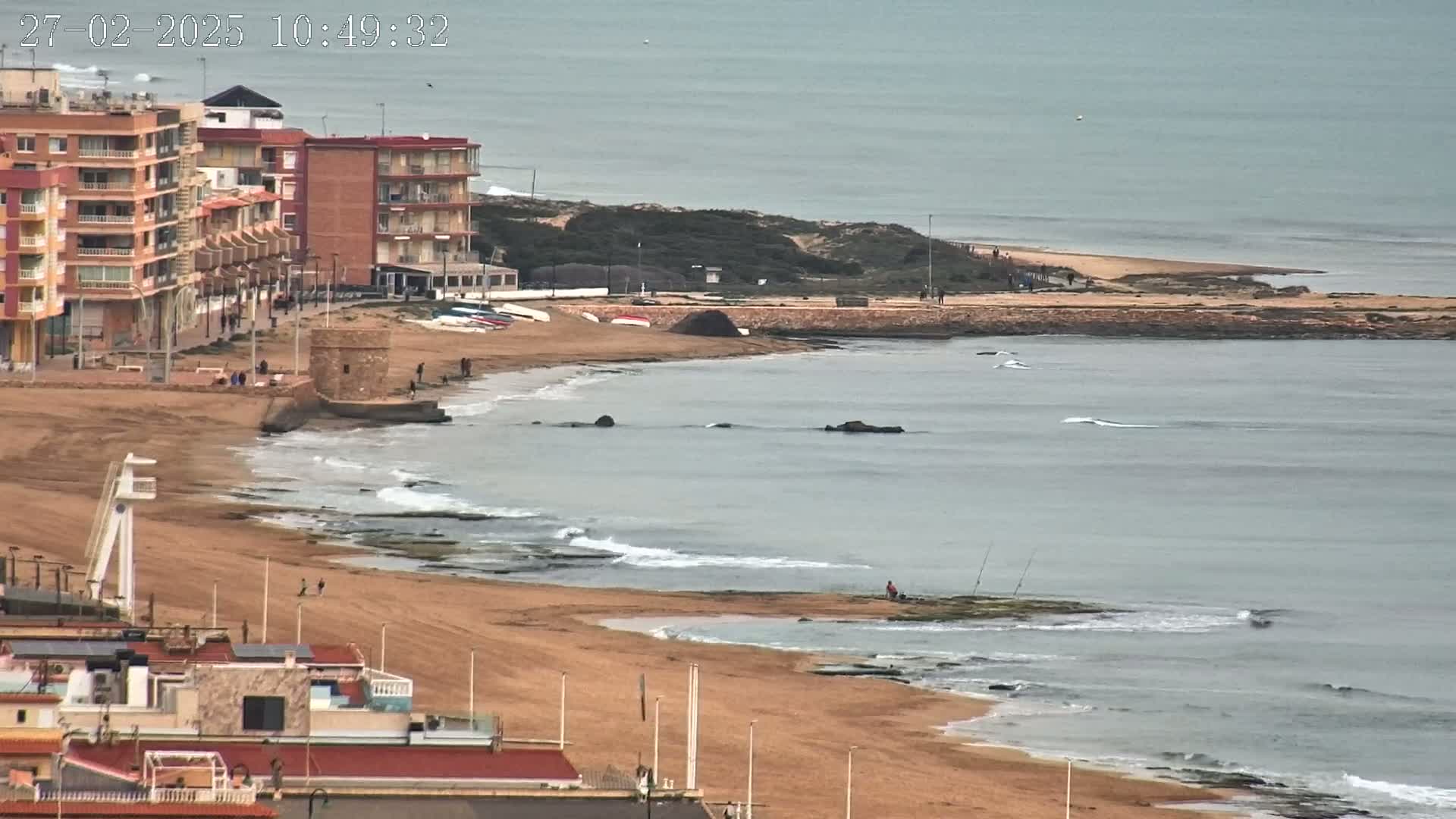 An overcast day shows a sandy beach with gentle waves lapping at the shore, buildings lining the coast, and a few people scattered along the beach and in the water.