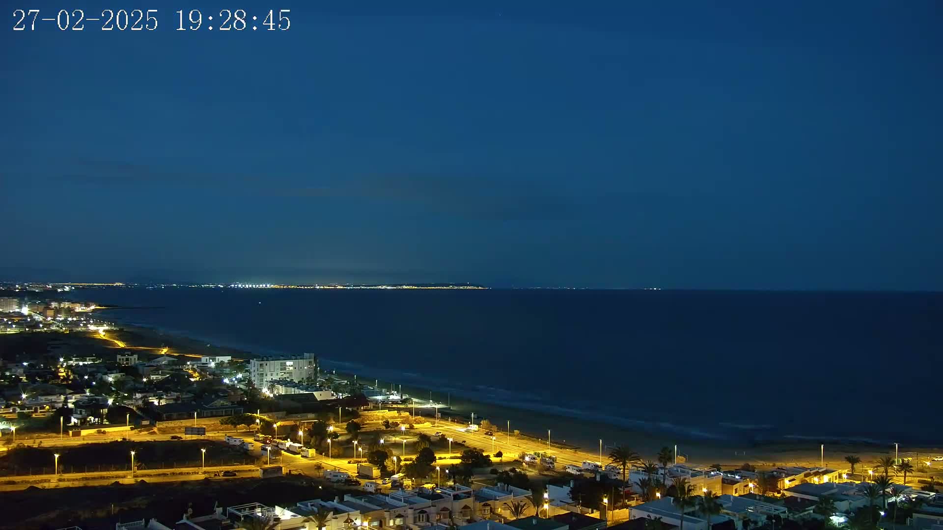 A nighttime coastal view shows a dark blue sea meeting a sandy beach lined with illuminated buildings and streetlights.