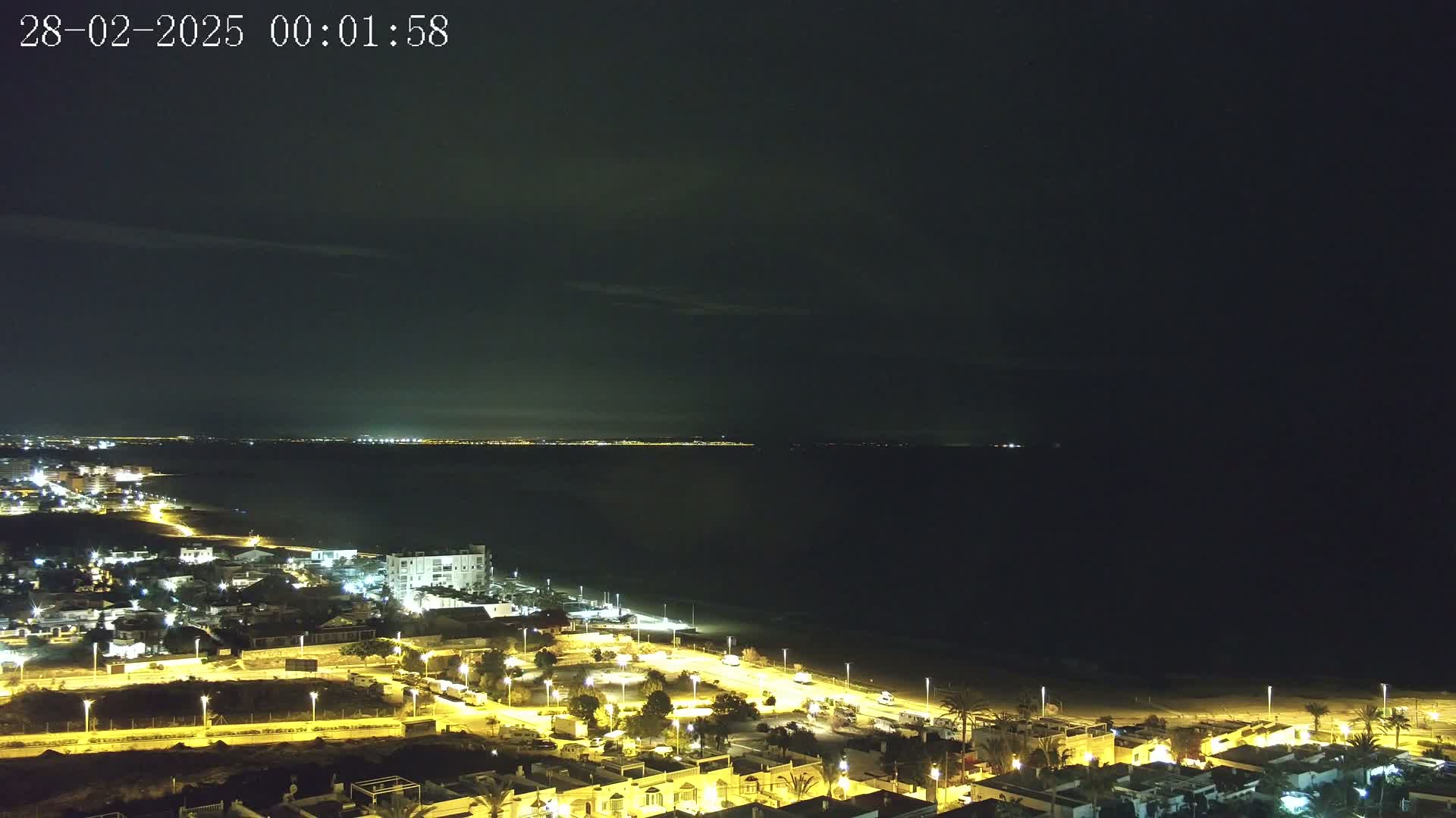 A nighttime coastal scene shows a dark ocean under a mostly clear sky, with a brightly lit town situated along the beach.