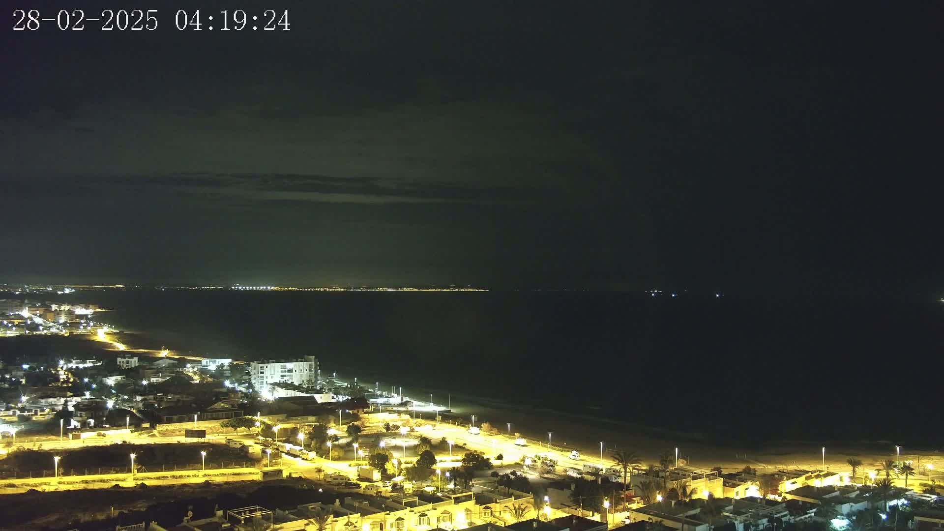 A nighttime coastal town is illuminated, with a dark ocean and a distant, faintly lit shoreline under a mostly clear, dark sky.