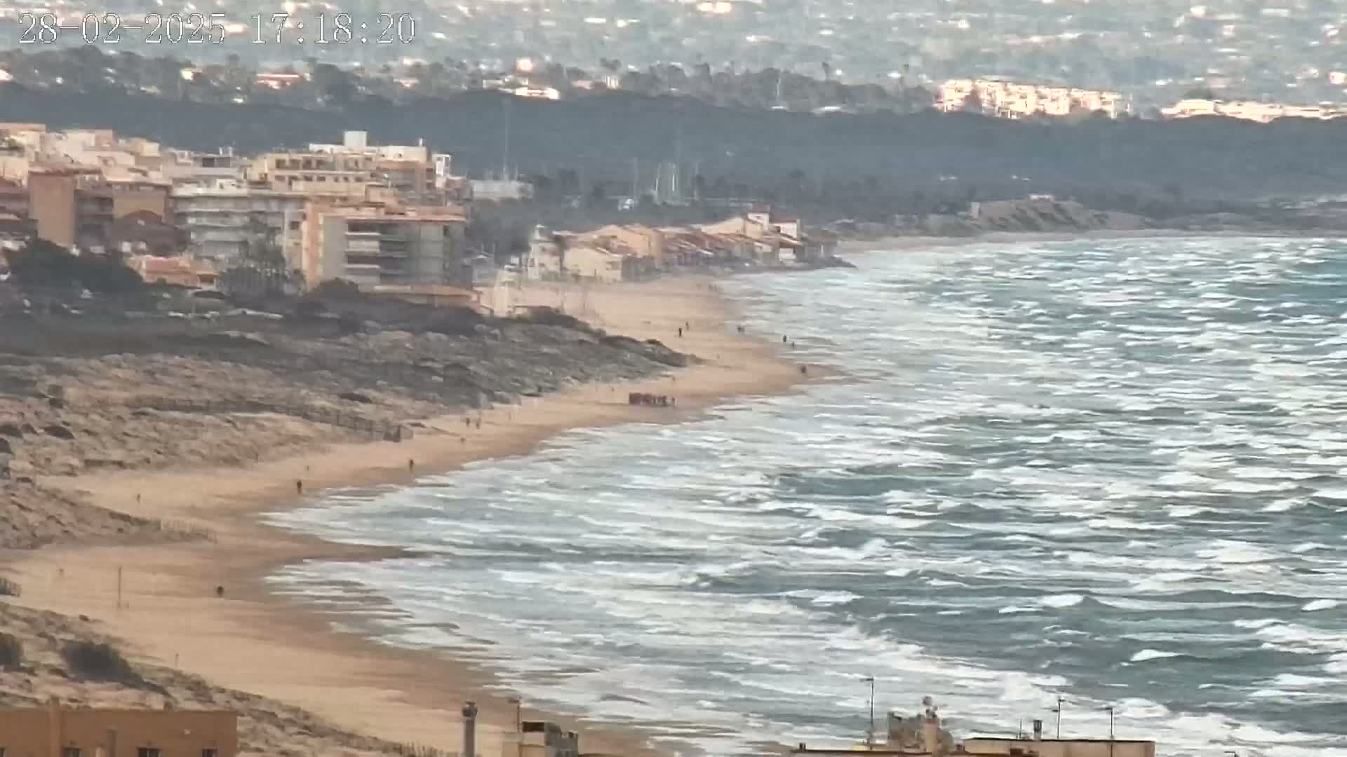 A long sandy beach with several people walking along it, bordered by buildings and a rocky coastline, is shown with choppy waves in a windy ocean.