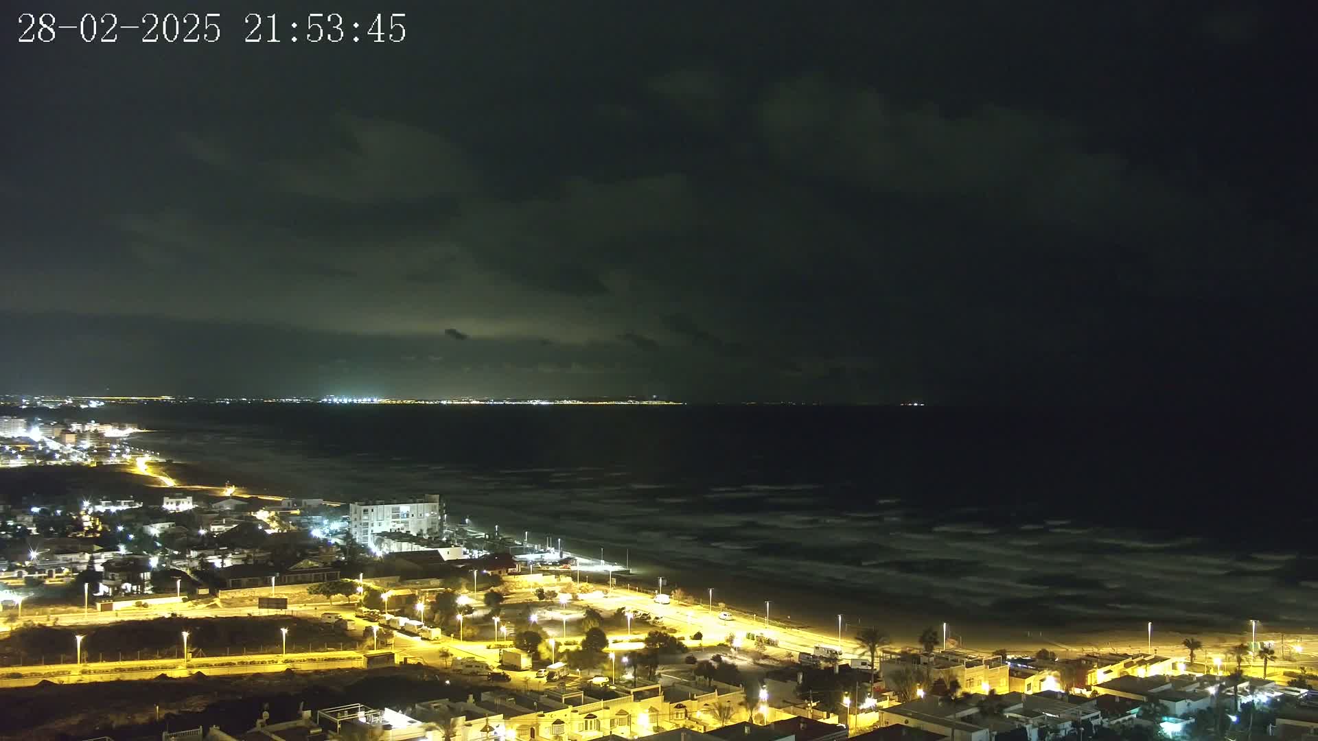 A nighttime coastal scene shows a dark, cloudy sky over a beach with gently waving water, and a town with illuminated buildings stretching into the distance.