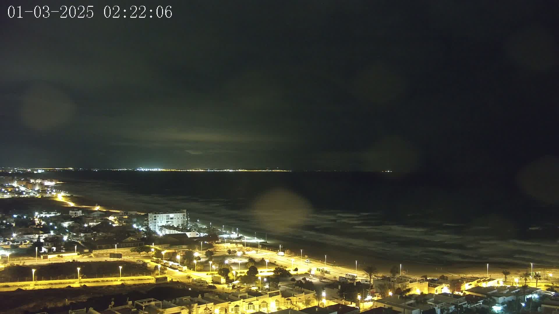 A nighttime coastal town illuminated by streetlights overlooks a dark, wavy ocean under a cloudy sky.