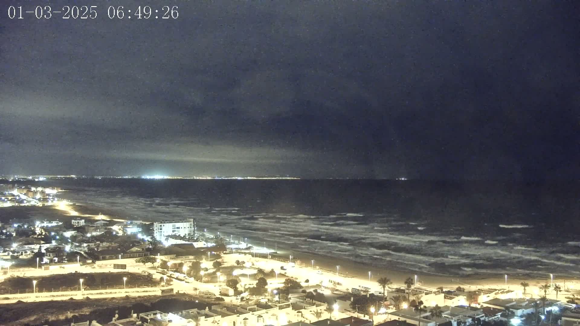 A nighttime coastal scene shows a dark, cloudy sky over a wavy ocean and a beach lined with illuminated buildings.