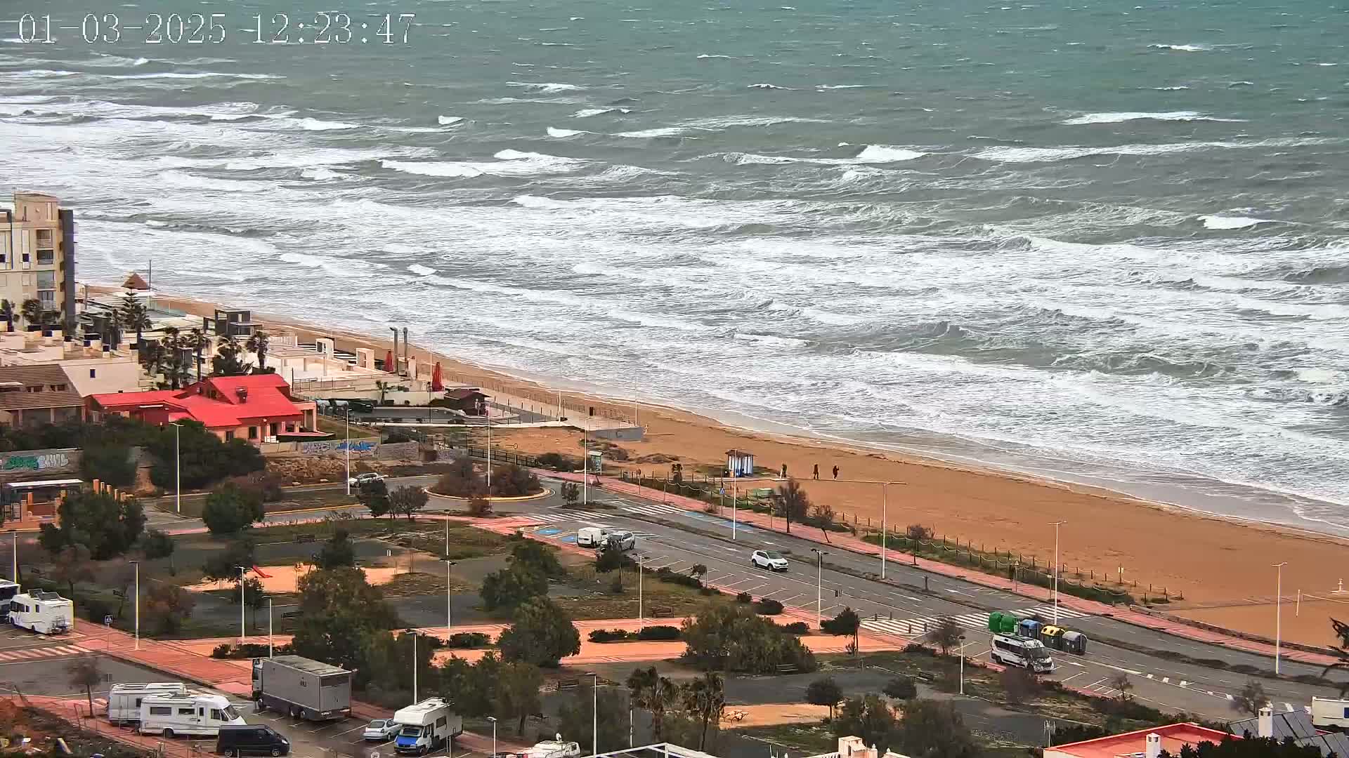 A windy day shows choppy waves crashing on a sandy beach next to a coastal road with parked cars and RVs.