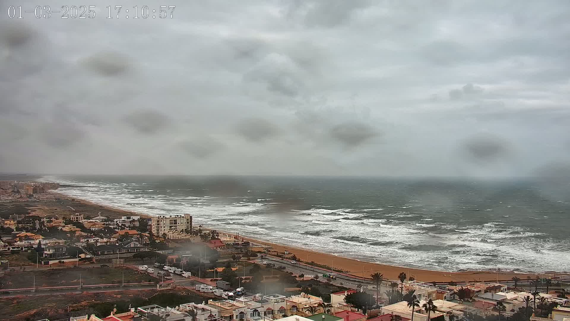 A coastal town is viewed from above on a cloudy, windy day with choppy waves crashing on the sandy shore.