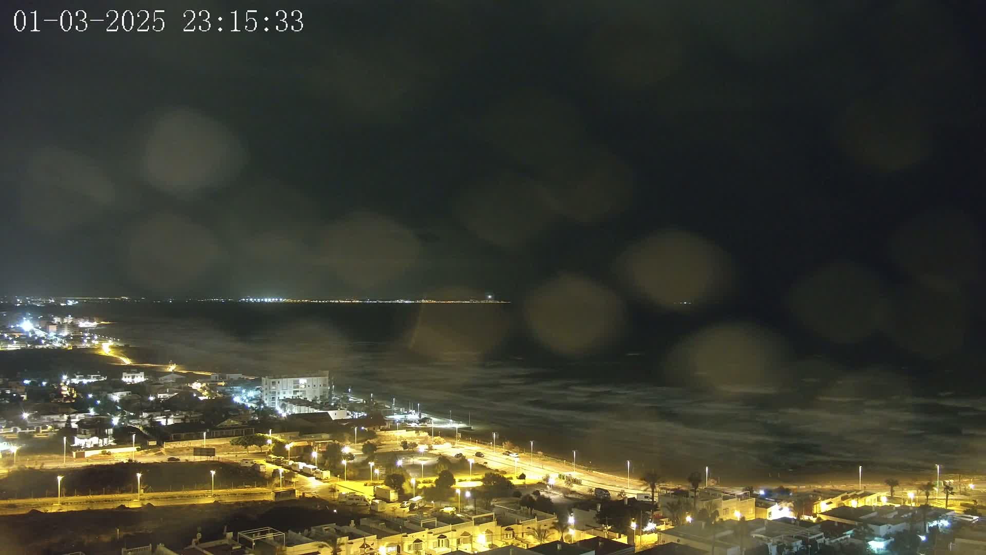A rain-streaked nighttime view shows a dark, wavy ocean meeting a brightly lit coastal town with a distant shoreline visible across the water.
