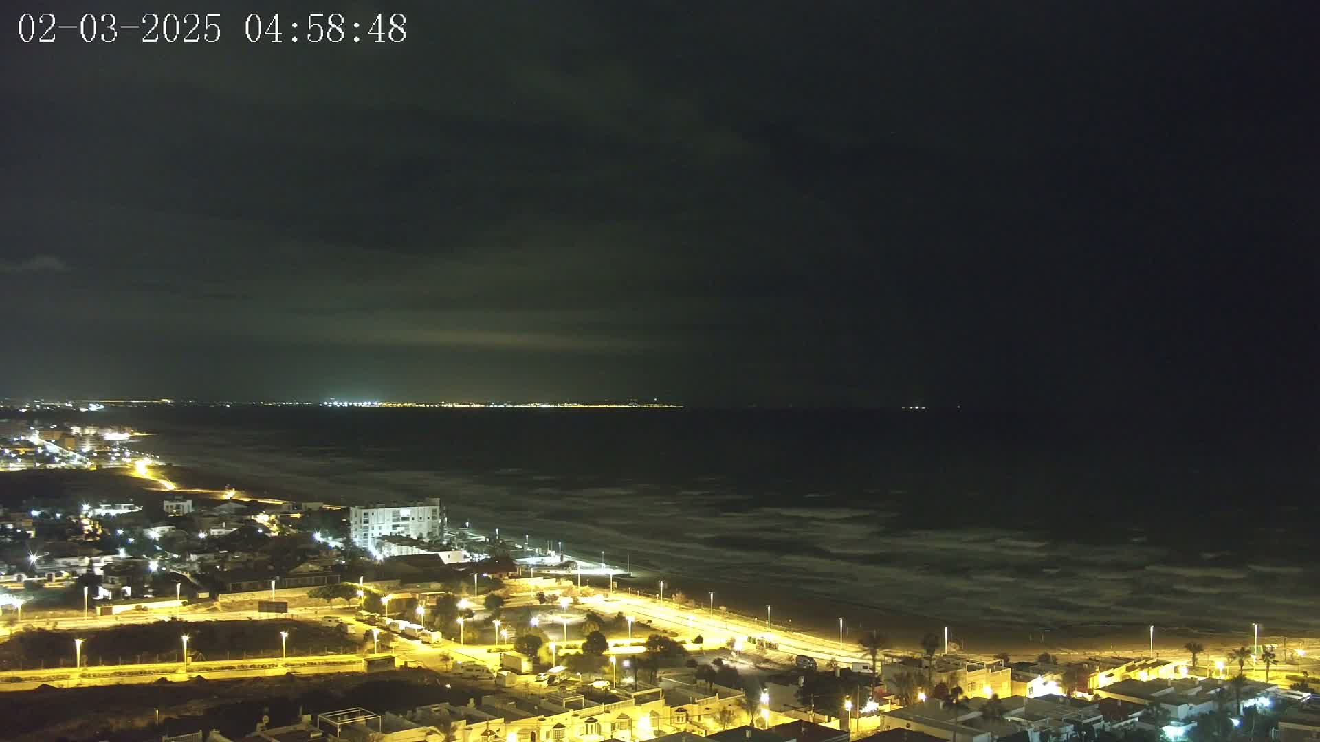 A night-time, high-angle view of a coastal town with illuminated streets and buildings overlooking a dark ocean with visible waves under a mostly cloudy sky.