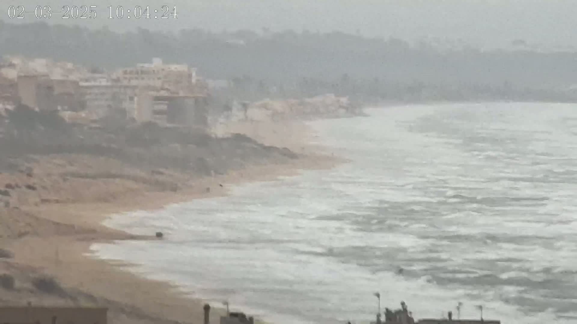 A hazy, overcast day reveals a sandy beach with choppy waves lapping against the shore near buildings on a cliff.