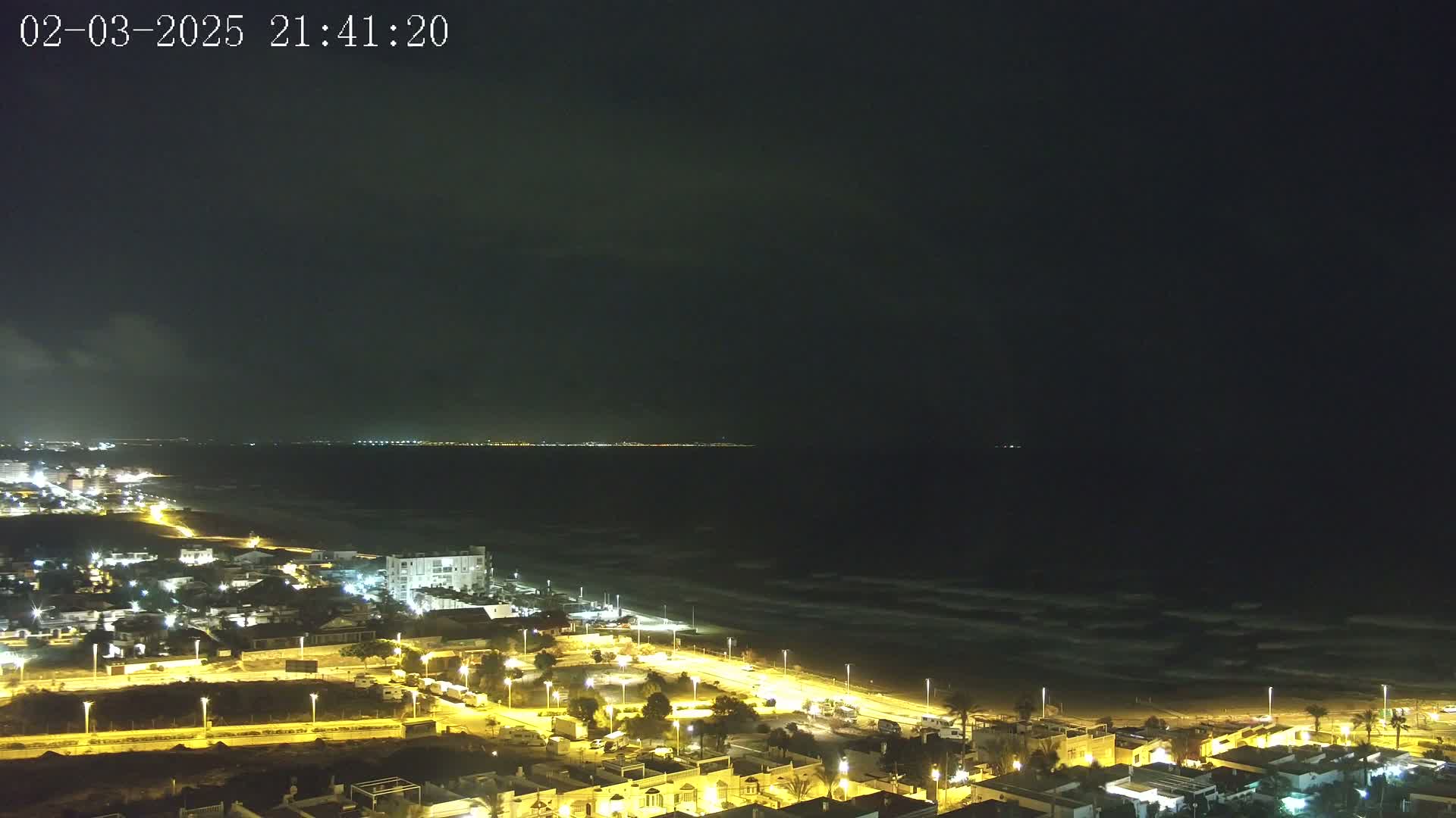 A nighttime coastal scene shows a dark ocean with small waves lapping a sandy beach, illuminated by streetlights along a residential area and a distant city skyline.