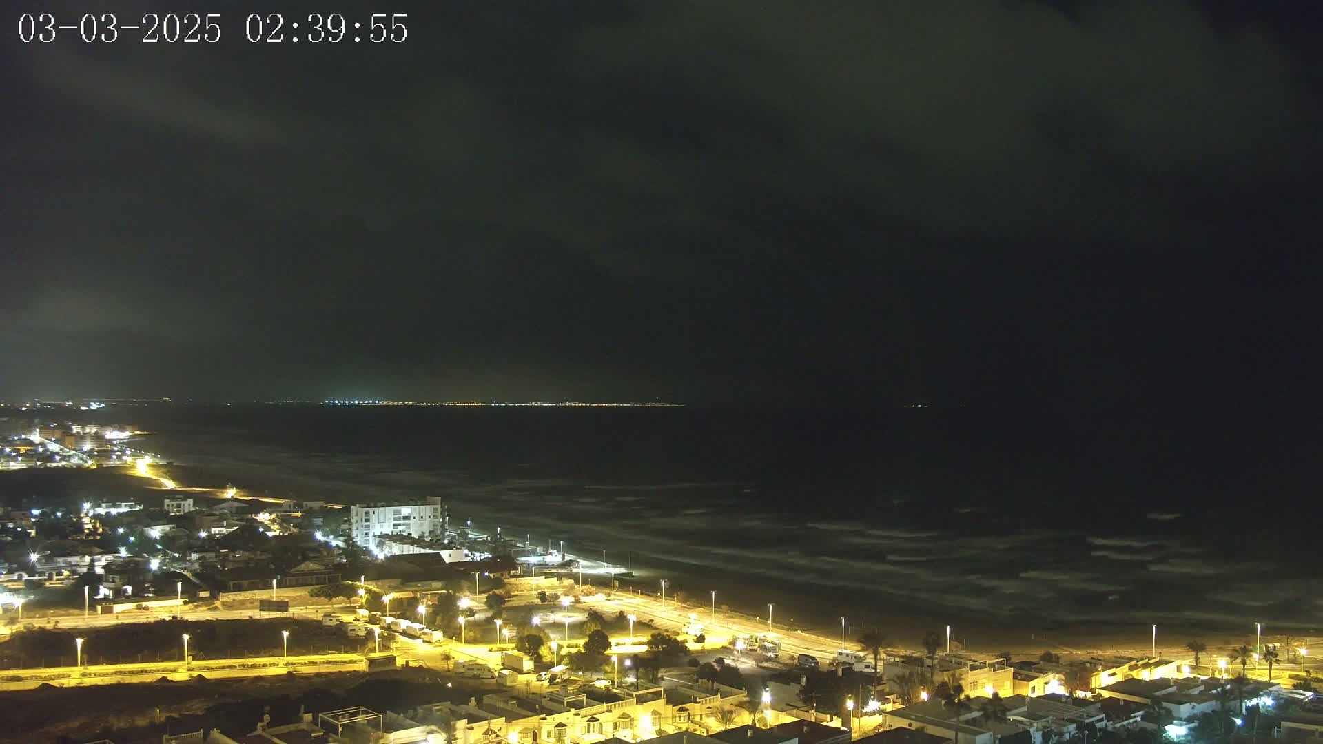 A nighttime coastal town with illuminated streets and buildings overlooks a dark ocean with visible waves under a cloudy sky.