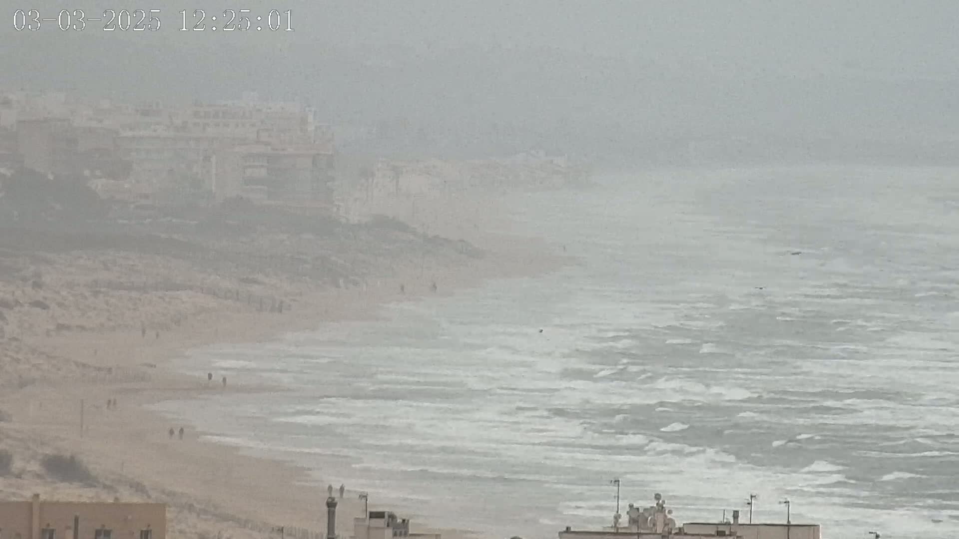 A hazy, overcast day shows a beach with a few people walking along the sand, and a choppy ocean with white capped waves, beyond which are distant buildings.