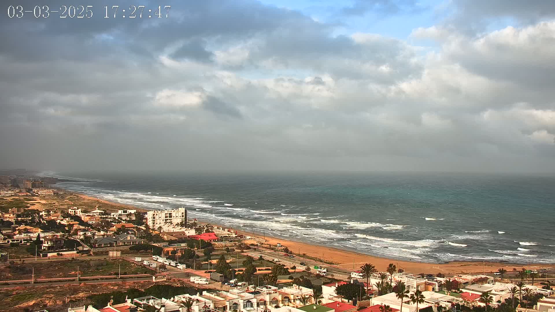 A coastal town with white buildings under a cloudy sky overlooks a beach with moderate waves and a somewhat choppy ocean.