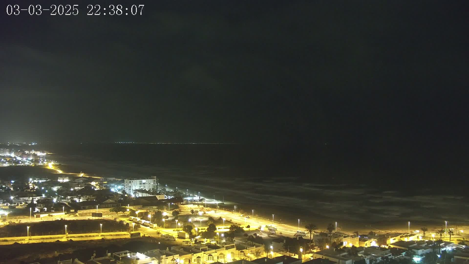 A nighttime aerial view shows a coastal town illuminated by streetlights, with a dark ocean exhibiting some wave activity under a mostly clear night sky.
