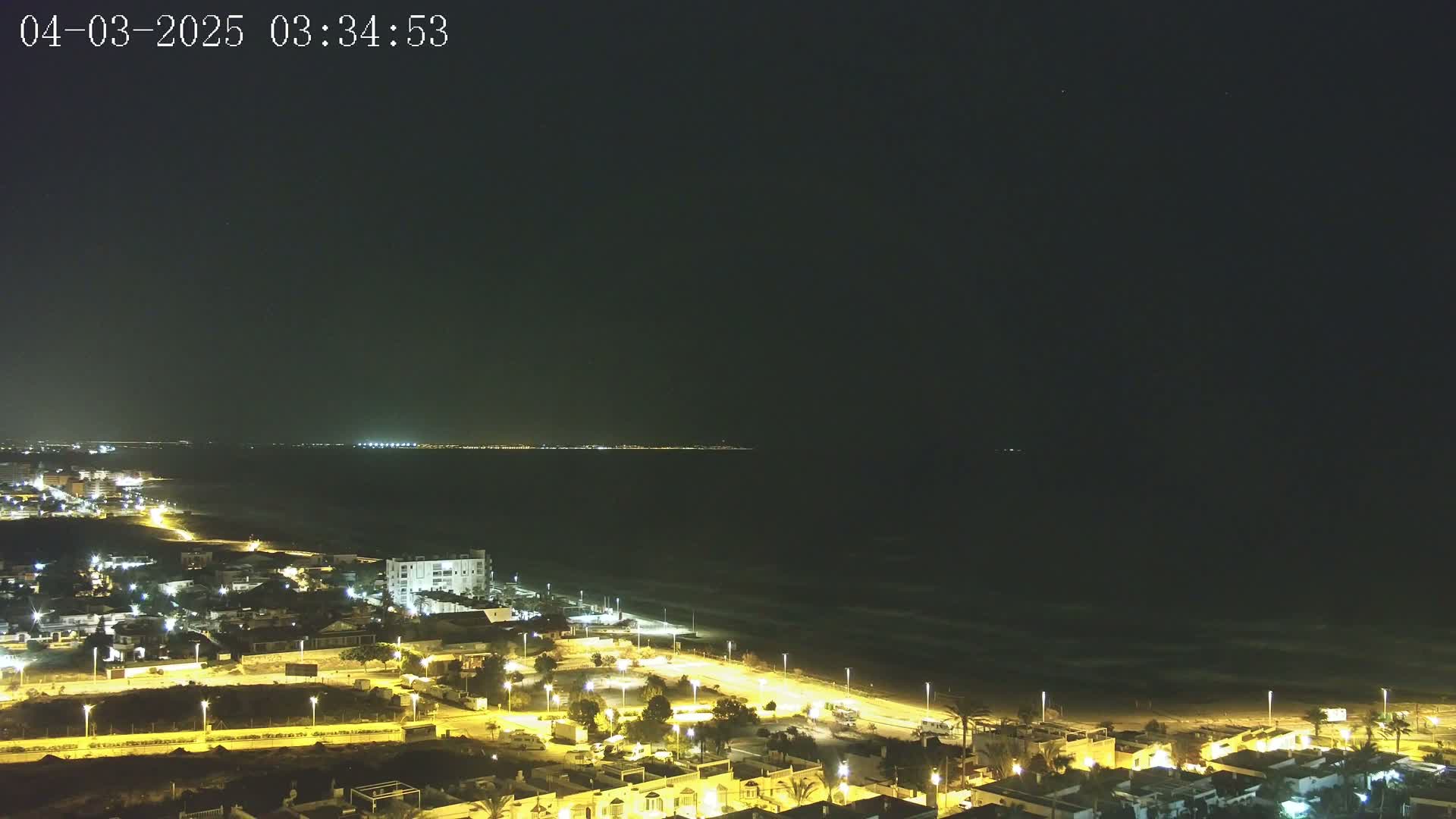 A nighttime, aerial view shows a coastal town illuminated with streetlights, extending to a dark ocean with gentle waves under a clear, dark sky.