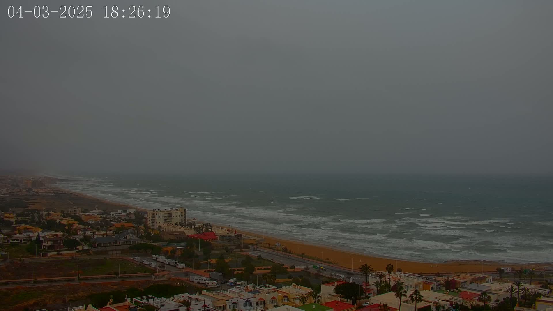 A coastal town is viewed from above on an overcast day, showing a beach with choppy waves under a gray sky.