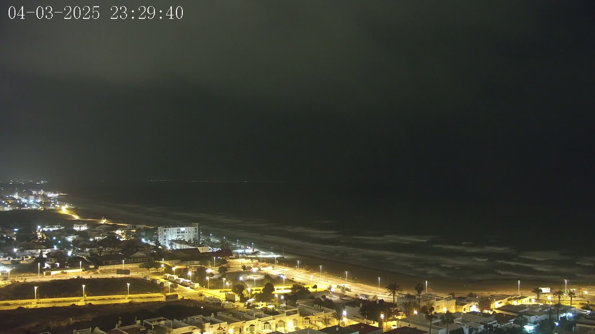 A nighttime coastal town is illuminated, with a dark, overcast sky and moderately wavy ocean visible in the background.