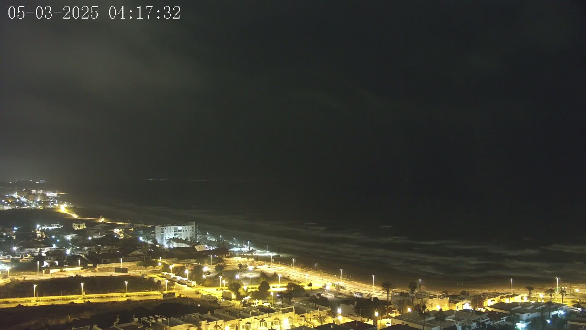 A nighttime, high-angle view of a coastal town illuminated with streetlights, next to a dark ocean with small waves under a mostly clear, dark sky.
