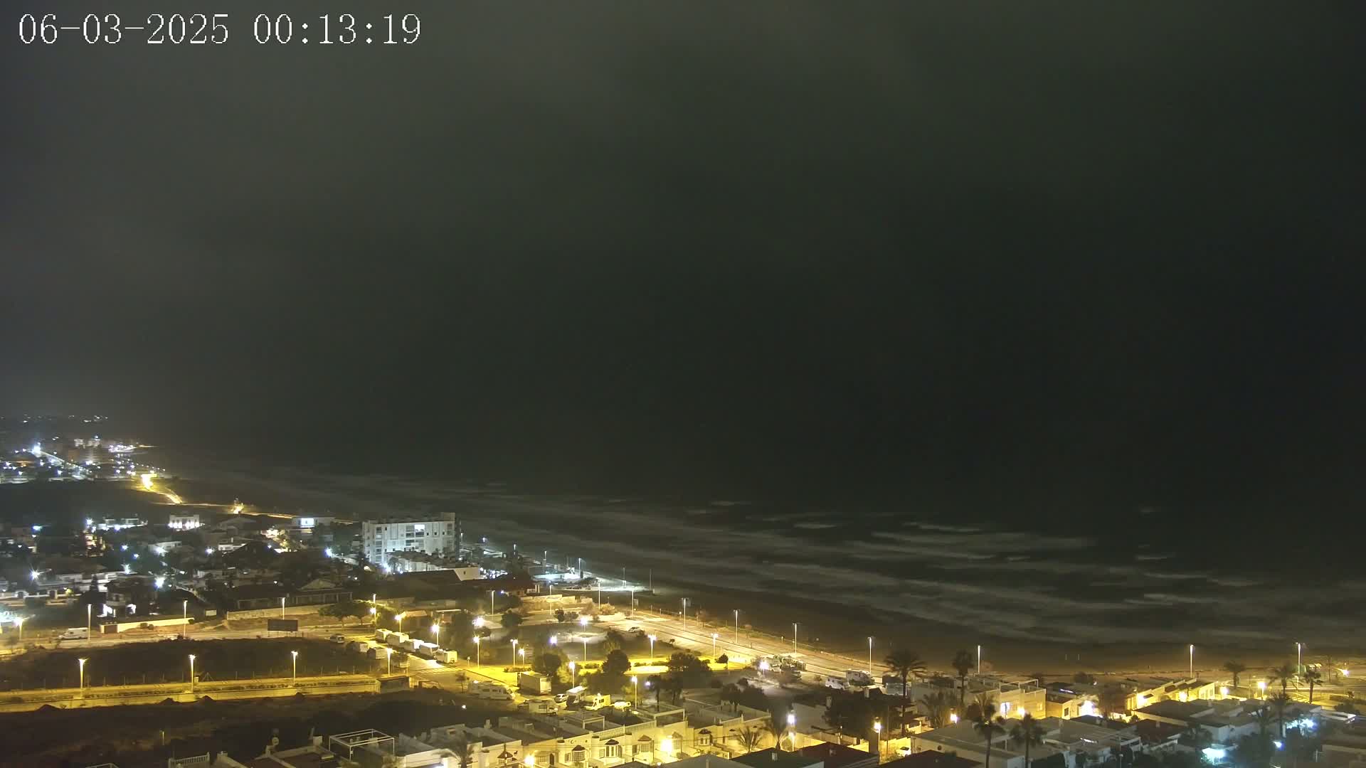 A nighttime coastal town with illuminated streets and buildings overlooks a dark, wavy ocean under a mostly cloudy sky.