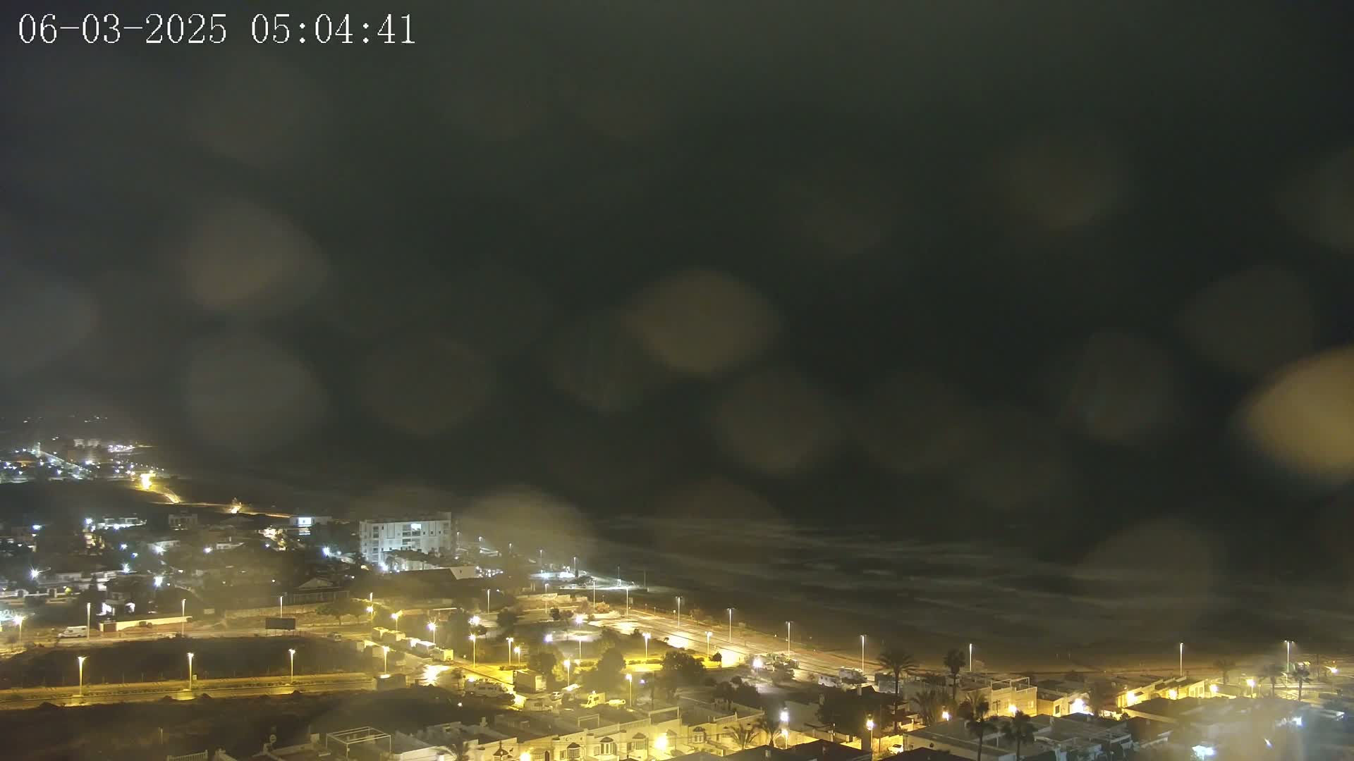 A nighttime coastal town is seen through rain-streaked glass, with the ocean visible in the background.