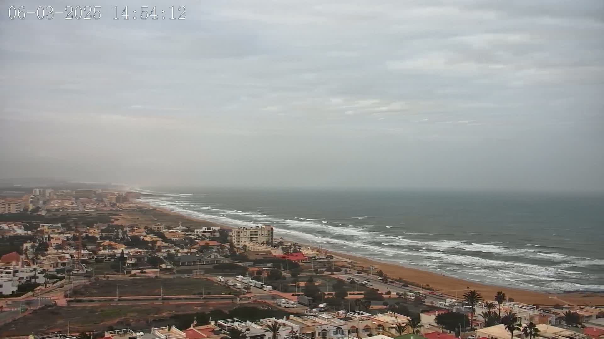 A hazy coastal town overlooks a beach with moderate waves under a cloudy sky.