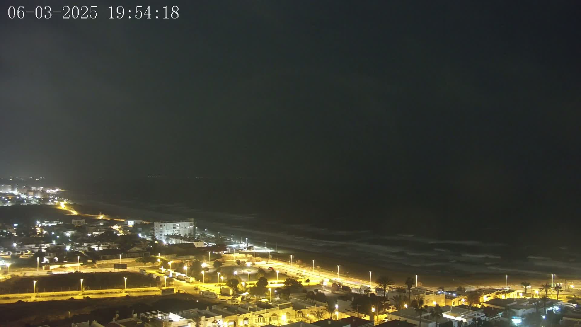 A nighttime aerial view shows a coastal town with illuminated streets and buildings next to a dark ocean with waves under a mostly clear, dark sky.