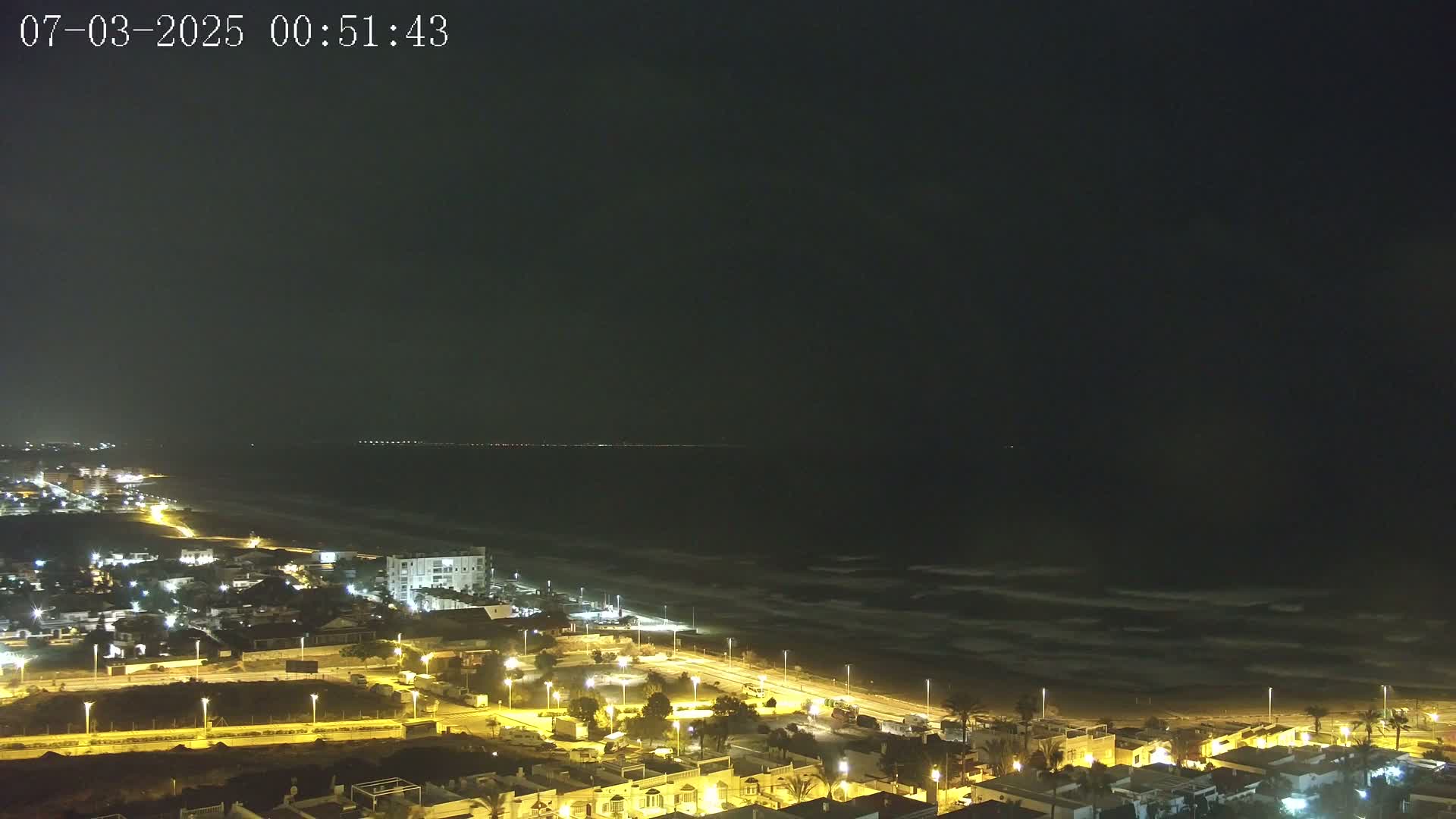 A nighttime, aerial view of a coastal town with illuminated streets and buildings, overlooking a dark ocean with some waves under a dark sky.