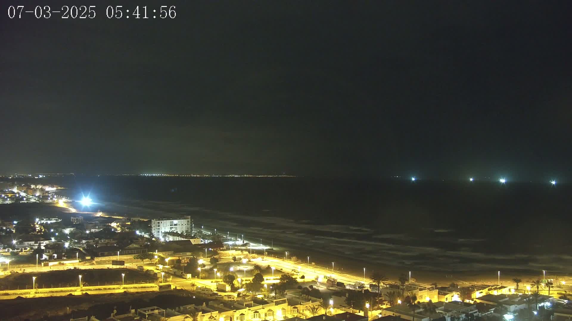 A nighttime coastal scene shows a dark ocean with small waves under a mostly clear sky, overlooking a town with lit streets and buildings along a sandy beach.