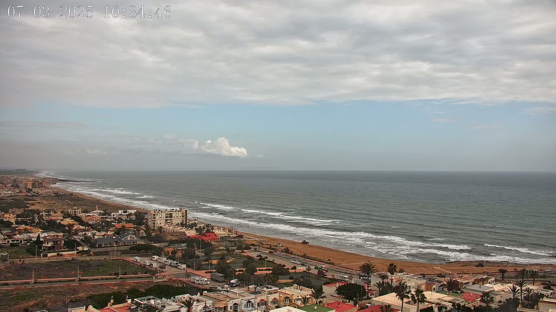 An expansive view shows a sandy beach with gentle waves meeting a calm ocean under a mostly cloudy sky.