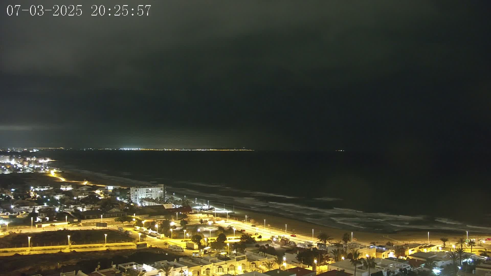A nighttime coastal scene shows a dark, overcast sky above a beach with gently lapping waves and a town with illuminated buildings.