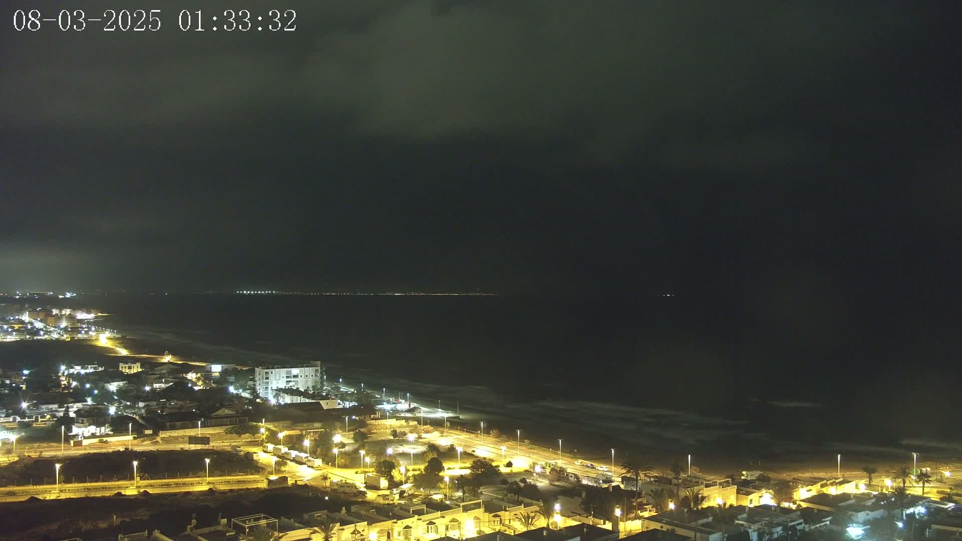 A nighttime coastal town is illuminated, with a dark, cloudy sky over a gently wavy ocean.