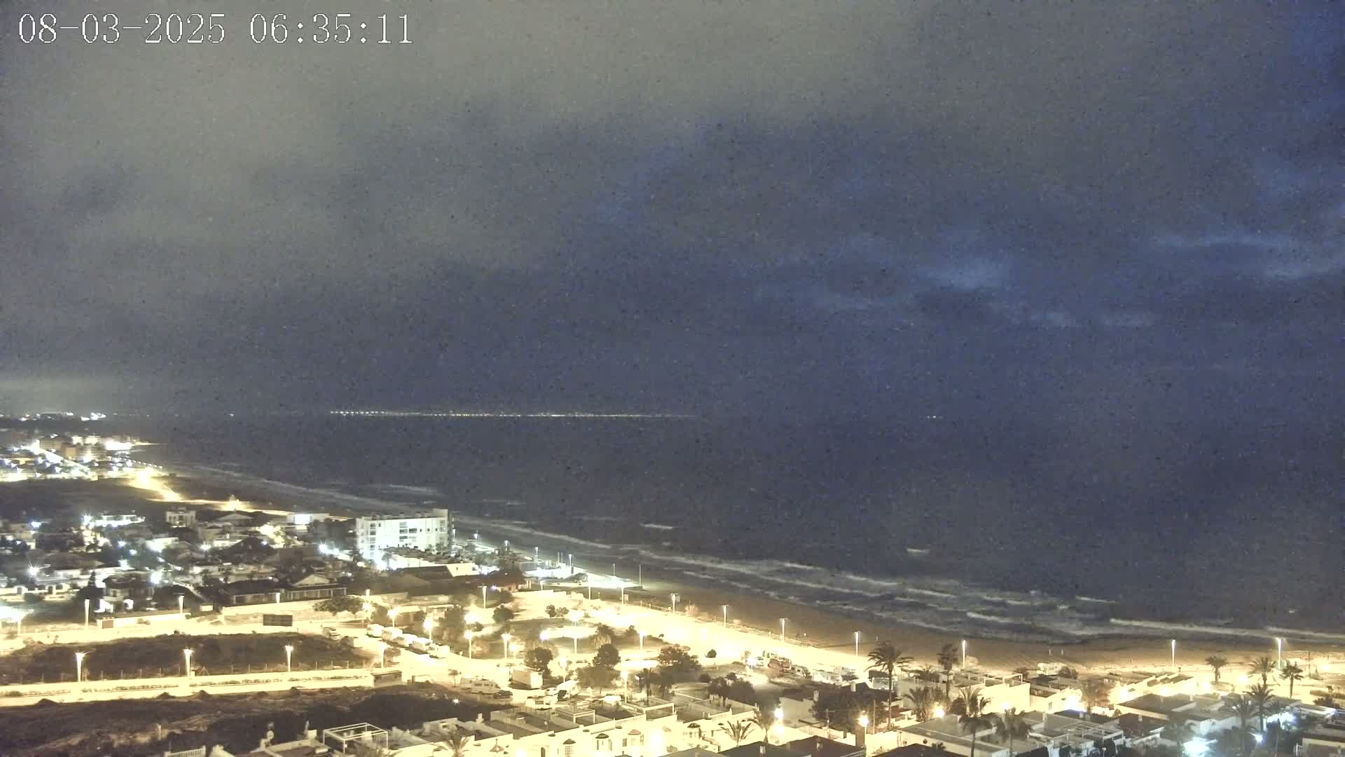 A nighttime view of a coastal town illuminated by streetlights, with a dark, overcast sky and waves gently lapping the shore.