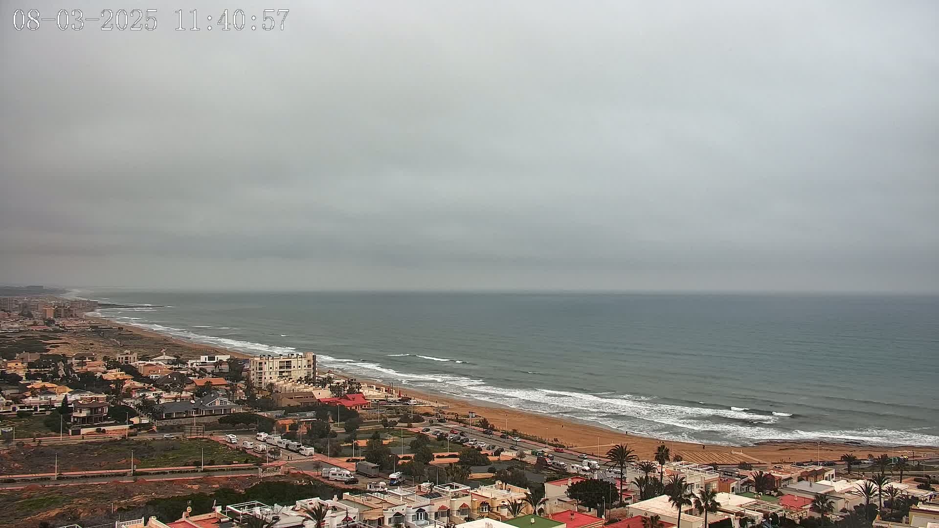 A coastal town sits under an overcast sky, overlooking a wide sandy beach and a calm sea with small waves.