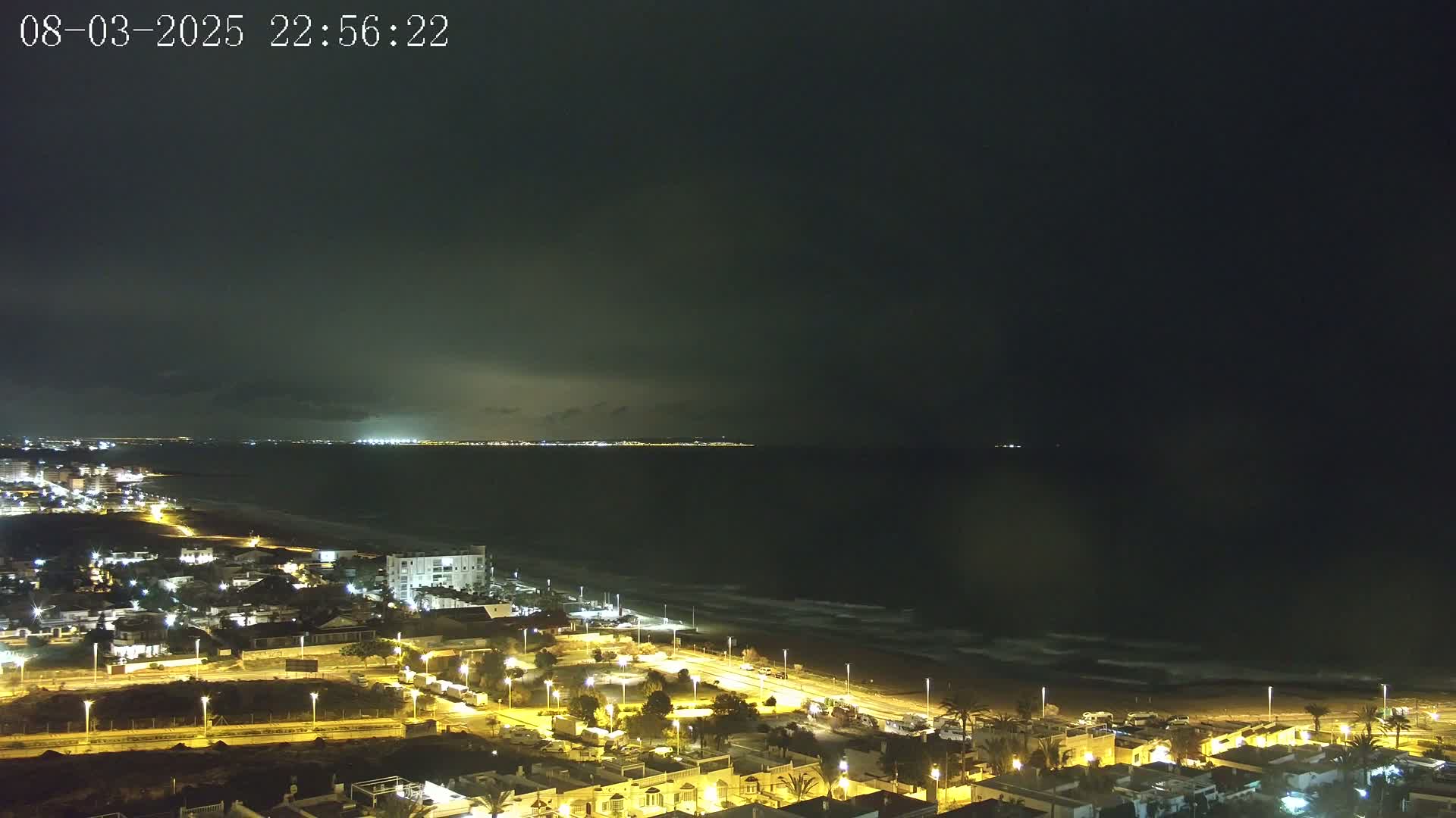 A nighttime coastal scene shows a dark, cloudy sky over a beach with gently rolling waves and a town illuminated by streetlights.