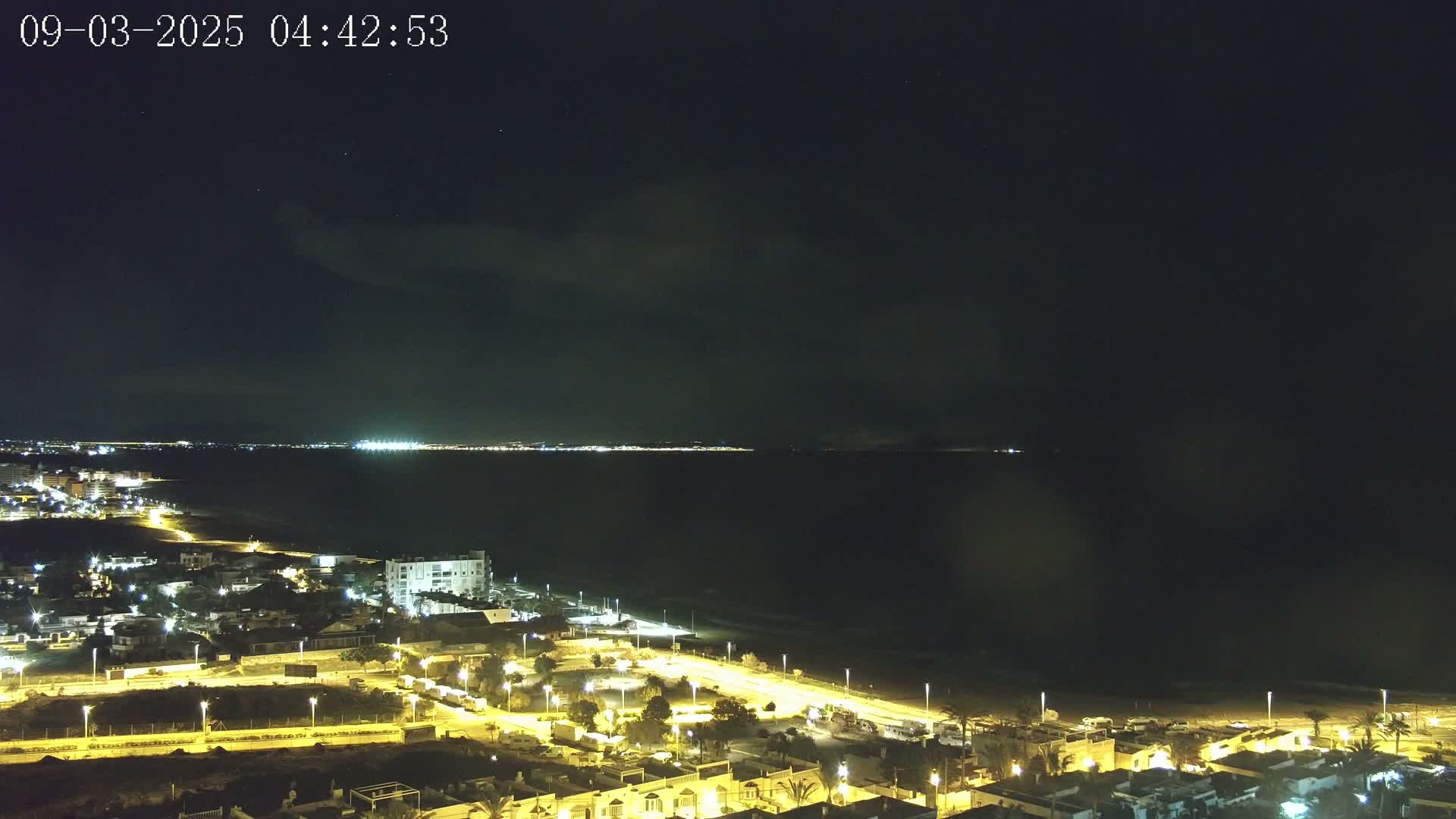 A nighttime coastal view shows a town's illuminated streets leading to a dark beach and calm sea, under a mostly clear, dark sky.
