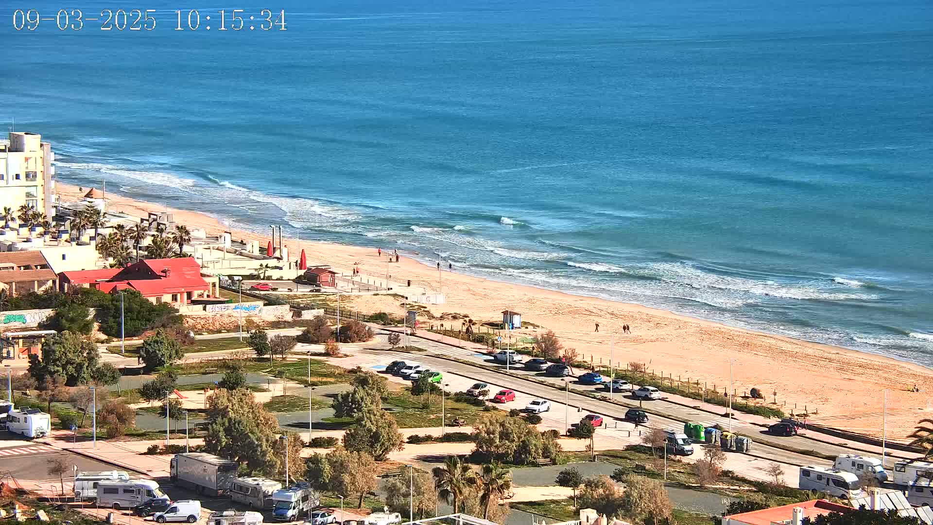 An aerial view shows a sandy beach with gently rolling waves on a sunny day, next to a parking lot filled with cars and recreational vehicles near buildings and greenery.