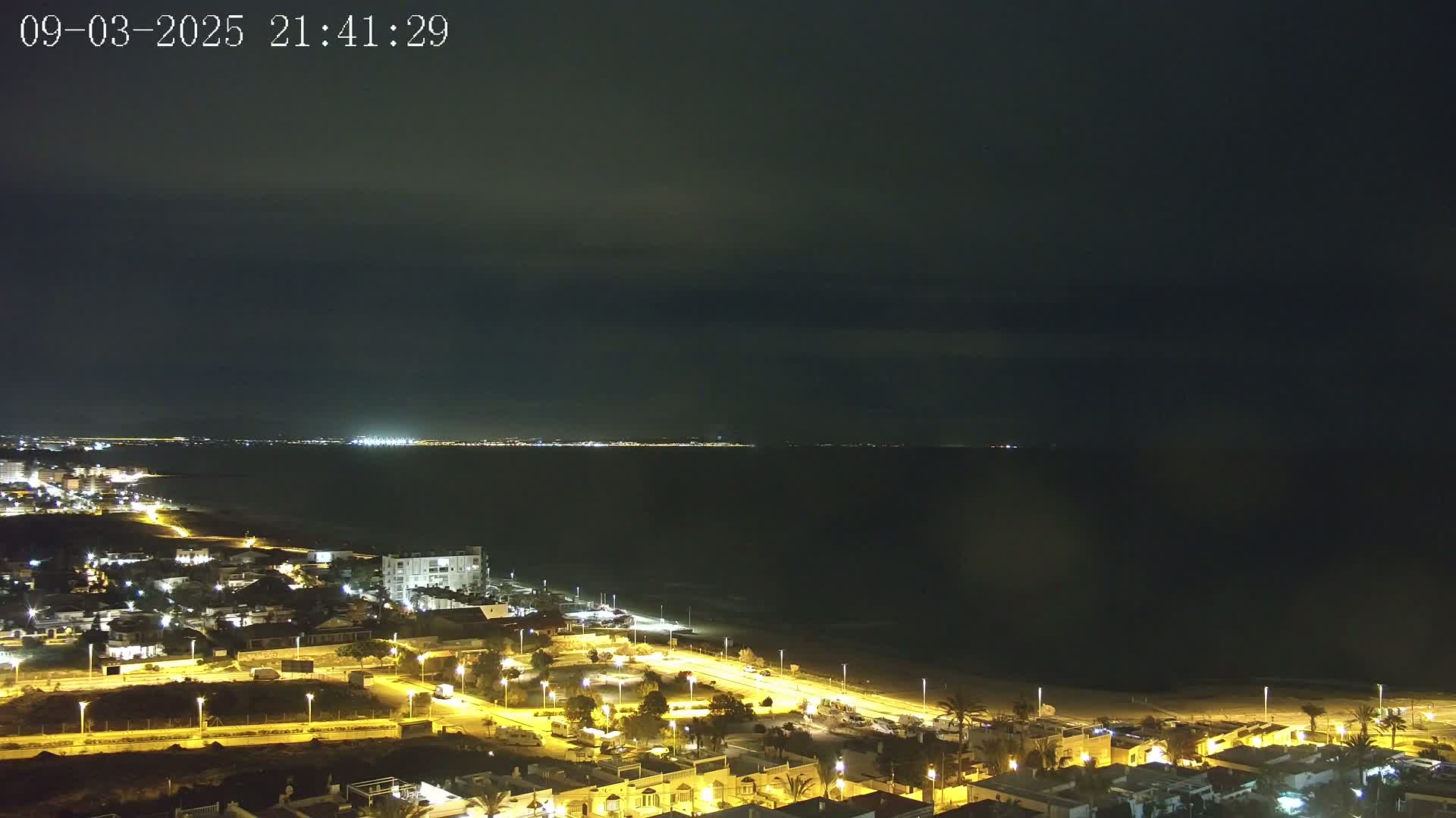 A nighttime coastal town illuminated by streetlights overlooks a dark sea under a mostly cloudy sky.