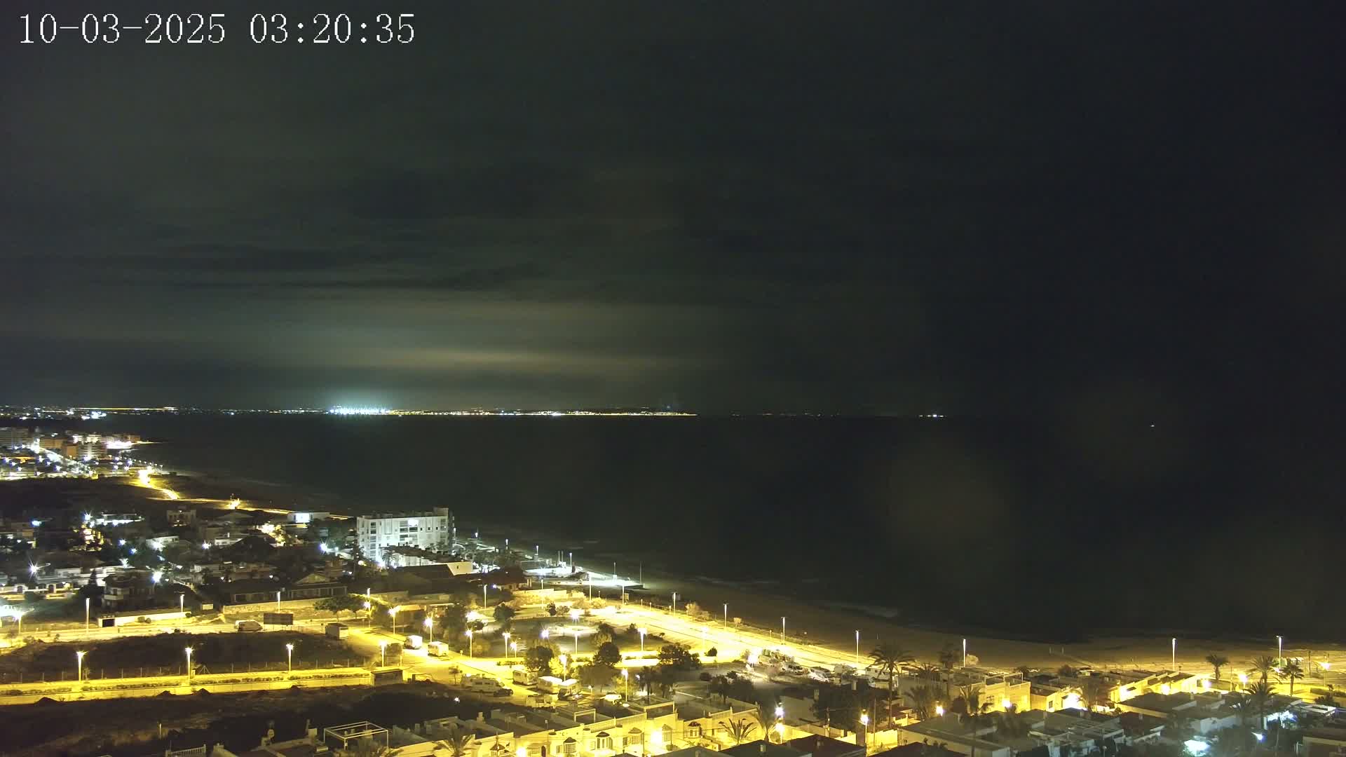 A nighttime coastal scene shows a dark ocean under a mostly cloudy sky, with a brightly lit town and beach in the foreground.