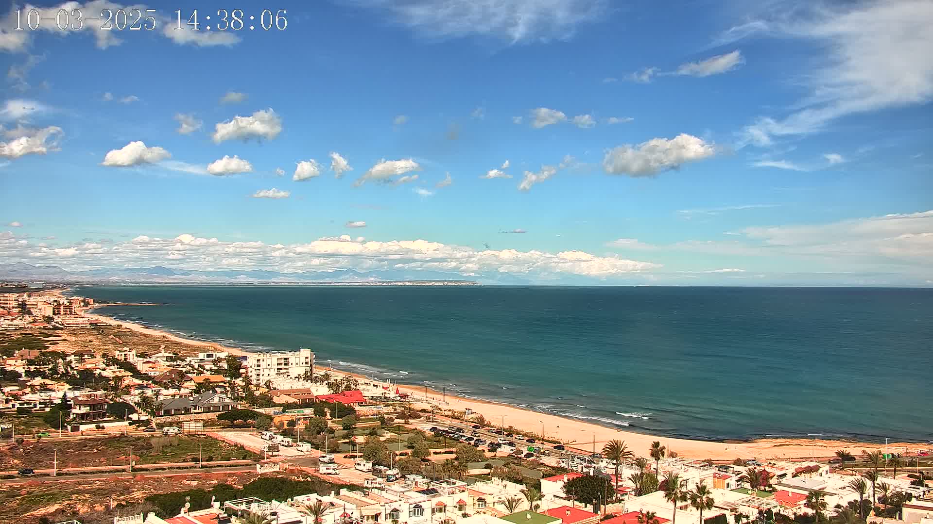 A coastal town with white buildings and palm trees sits beside a sandy beach and a calm, teal ocean under a mostly sunny sky with scattered cumulus clouds and distant mountains visible on the horizon.
