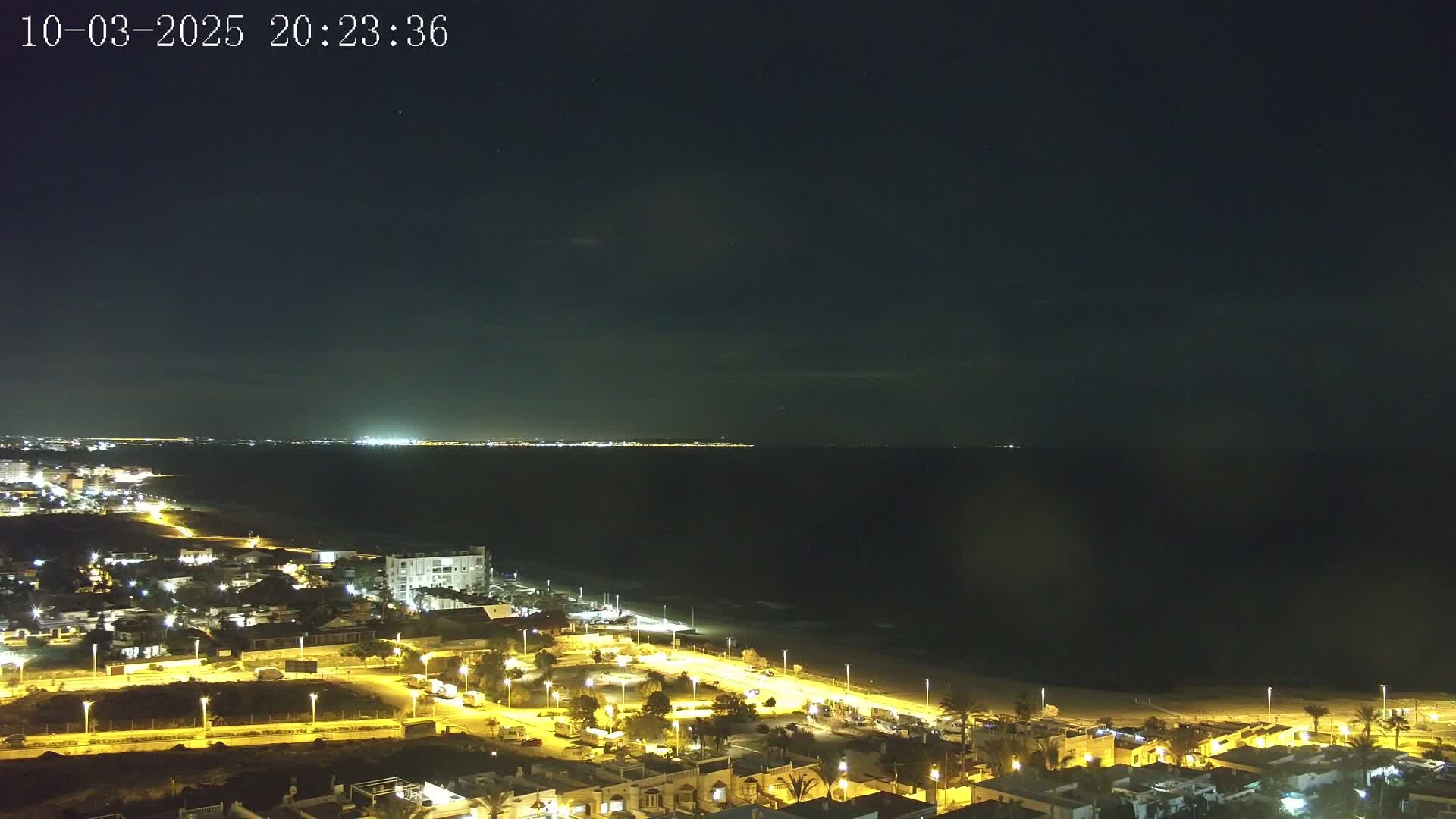 A nighttime coastal town is illuminated, with a dark ocean extending to a distant, faintly lit shoreline under a mostly clear sky.