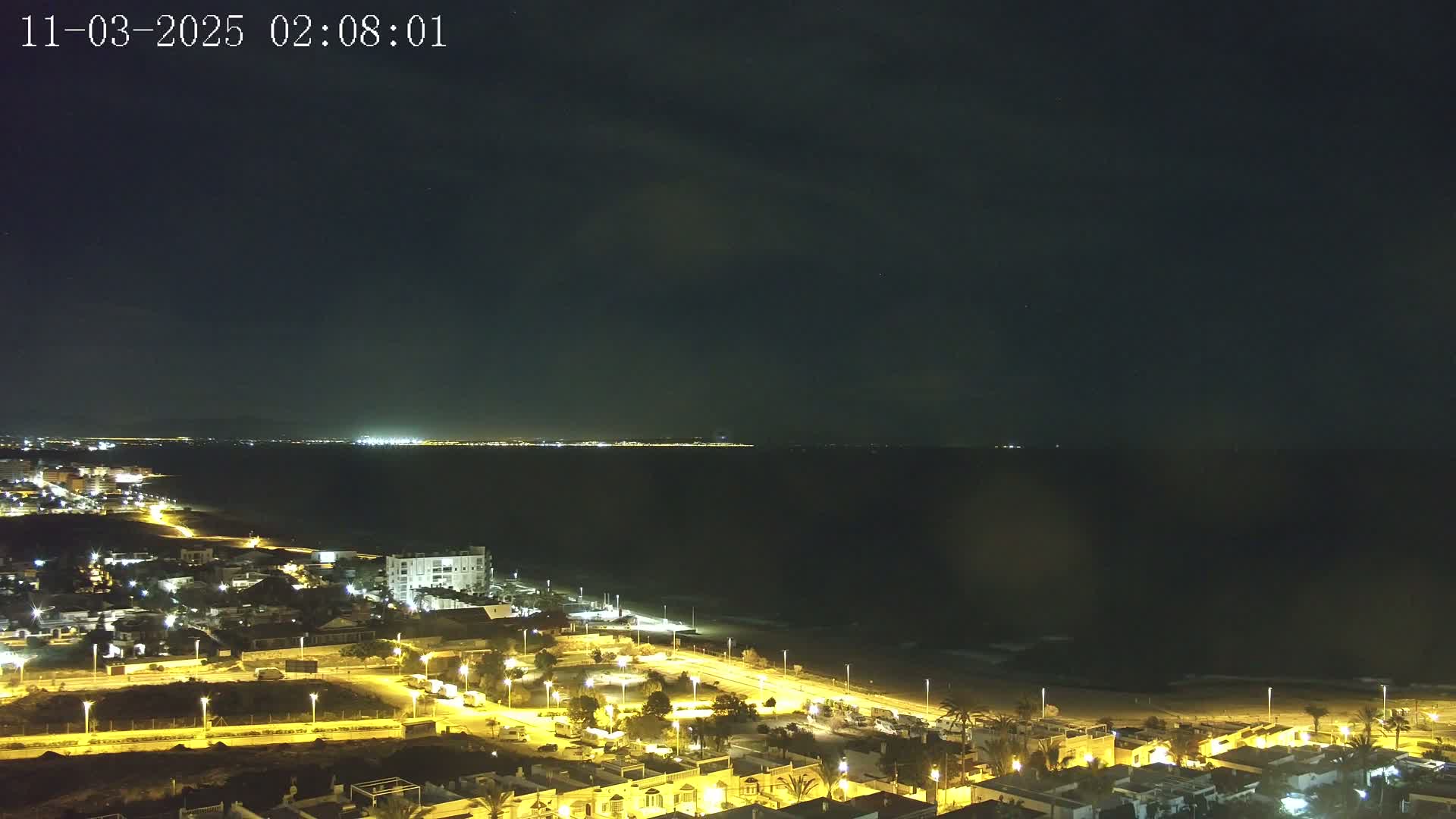 A nighttime coastal town is illuminated, with a dark ocean extending to a faintly lit distant shoreline under a mostly clear sky.