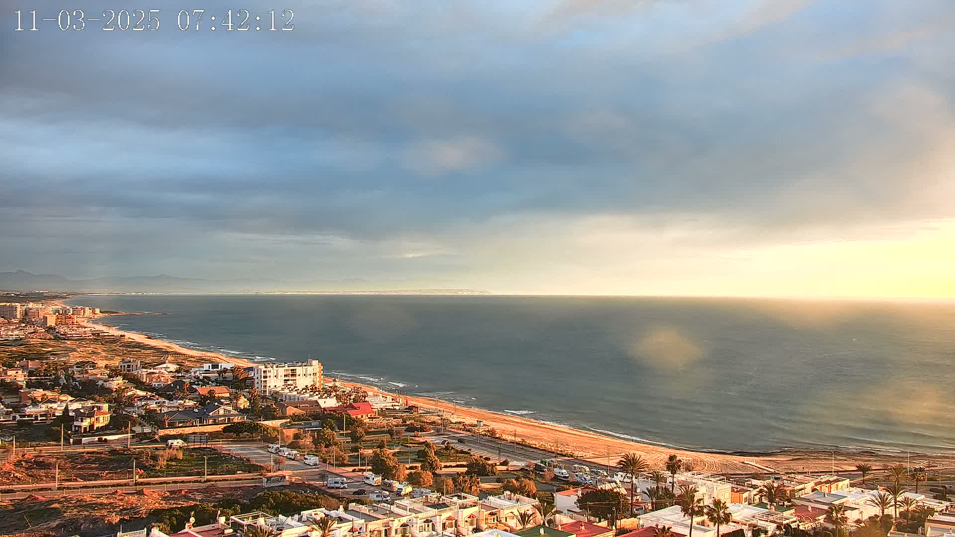 An elevated view of a coastal town on a partly cloudy morning shows a calm sea and sandy beach stretching to a distant mountain range.