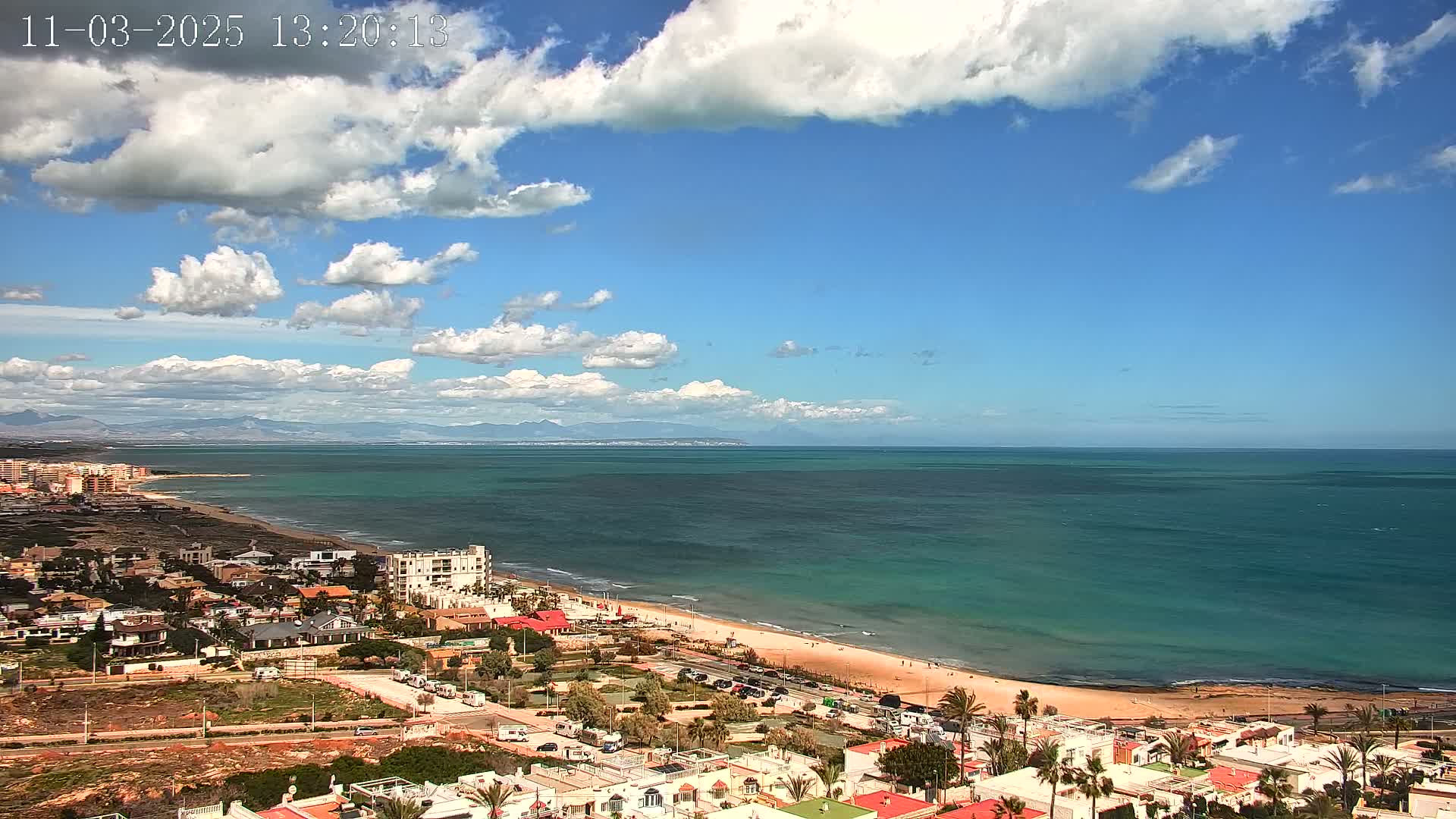 A coastal town with a sandy beach and turquoise ocean stretches to distant mountains under a partly cloudy blue sky.