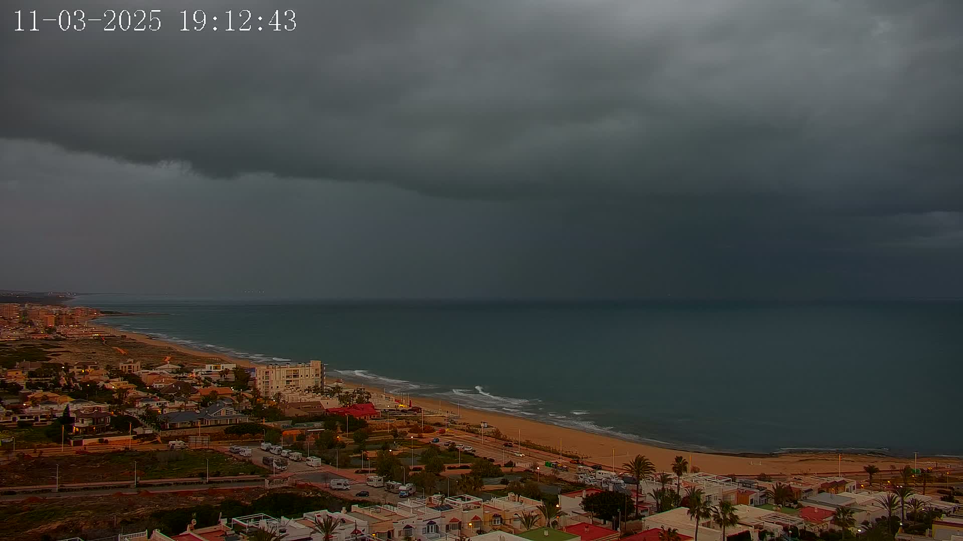 A coastal town sits under a dark, stormy sky, with a wide, sandy beach separating the town from a somewhat choppy sea.