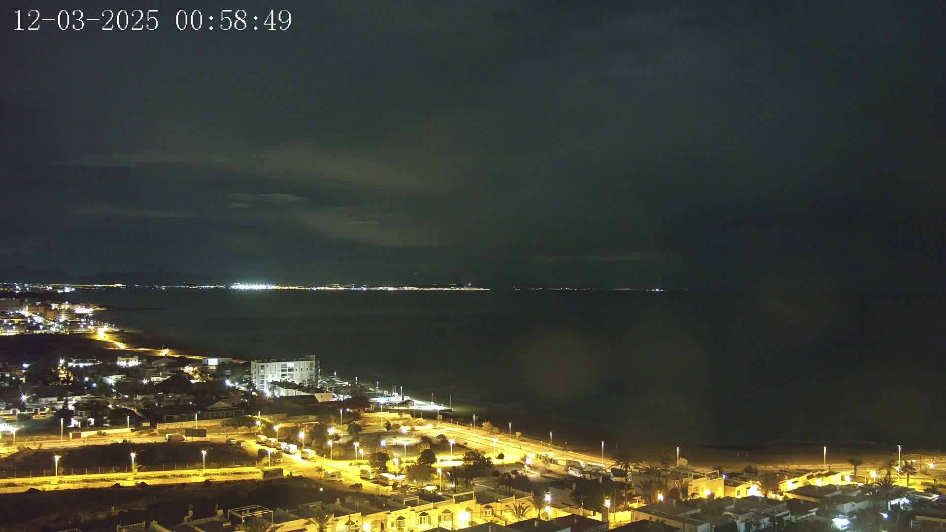 A nighttime view of a coastal town illuminated by streetlights, with a dark, cloudy sky over a calm sea and a distant, faintly lit shoreline.