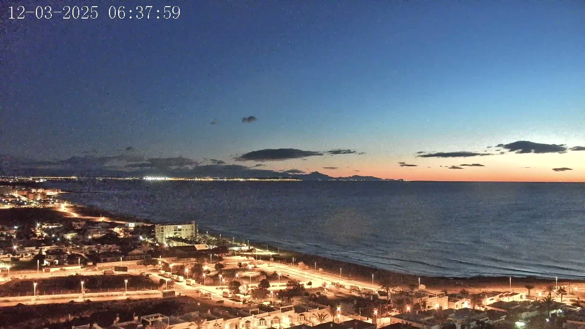 A coastal town at twilight, with lights illuminating the streets and buildings along a dark beach, overlooks a calm sea under a mostly clear, dusky sky.