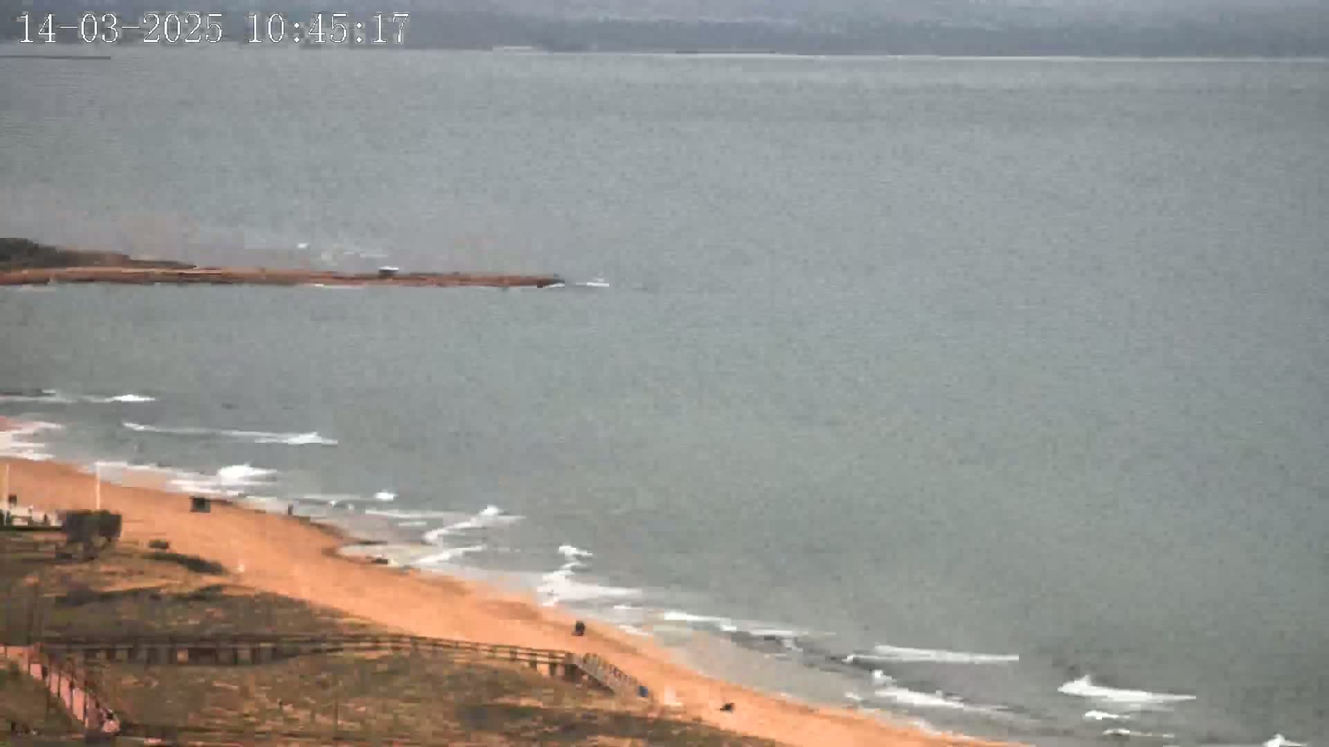 An overcast day shows a sandy beach with gentle waves lapping the shore, extending to a calm body of water.
